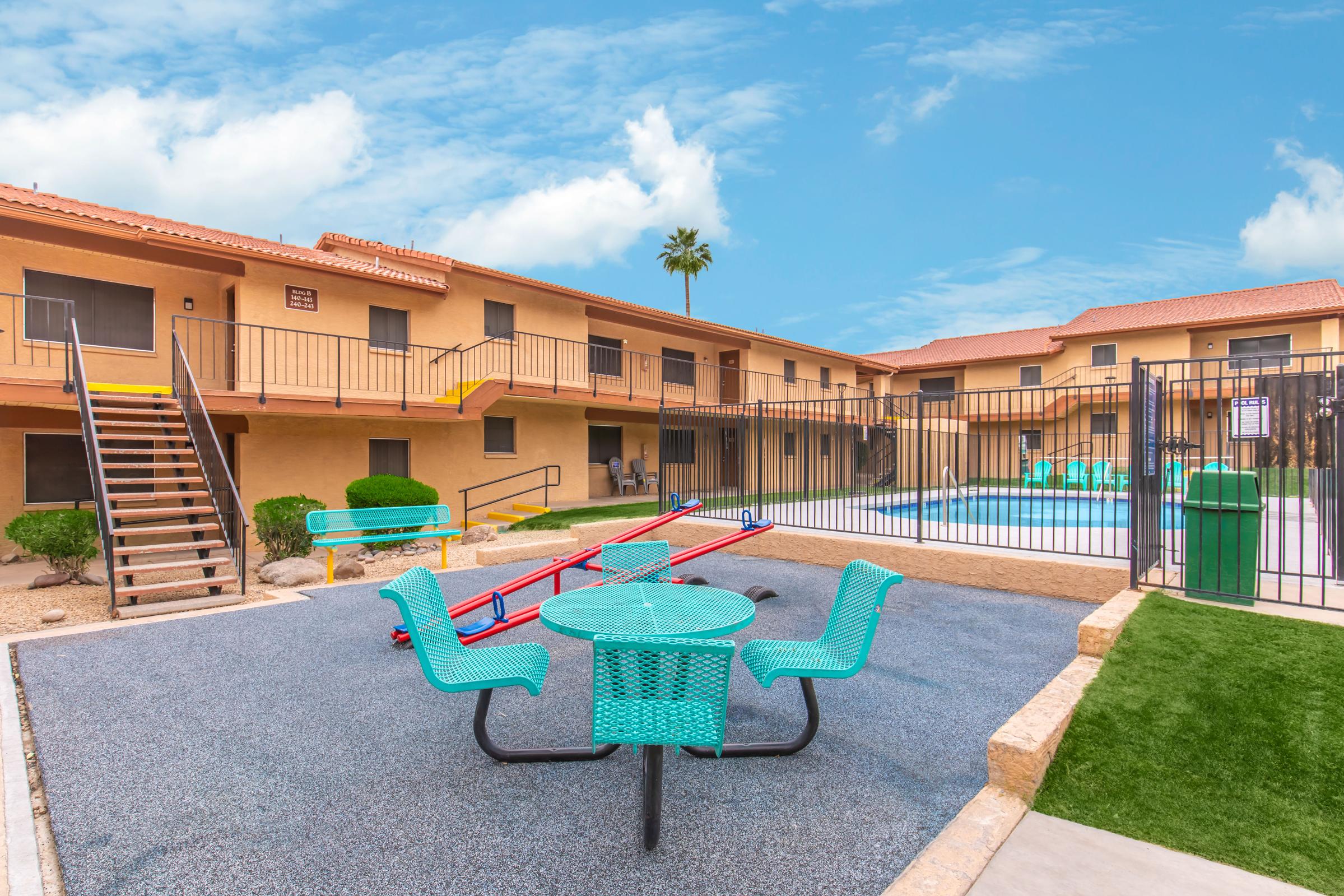 A view of a courtyard with a swimming pool surrounded by beige apartment buildings. Bright turquoise outdoor furniture, including a table and chairs, is placed on a textured surface. A seesaw is featured nearby, with palm trees and a clear blue sky in the background.