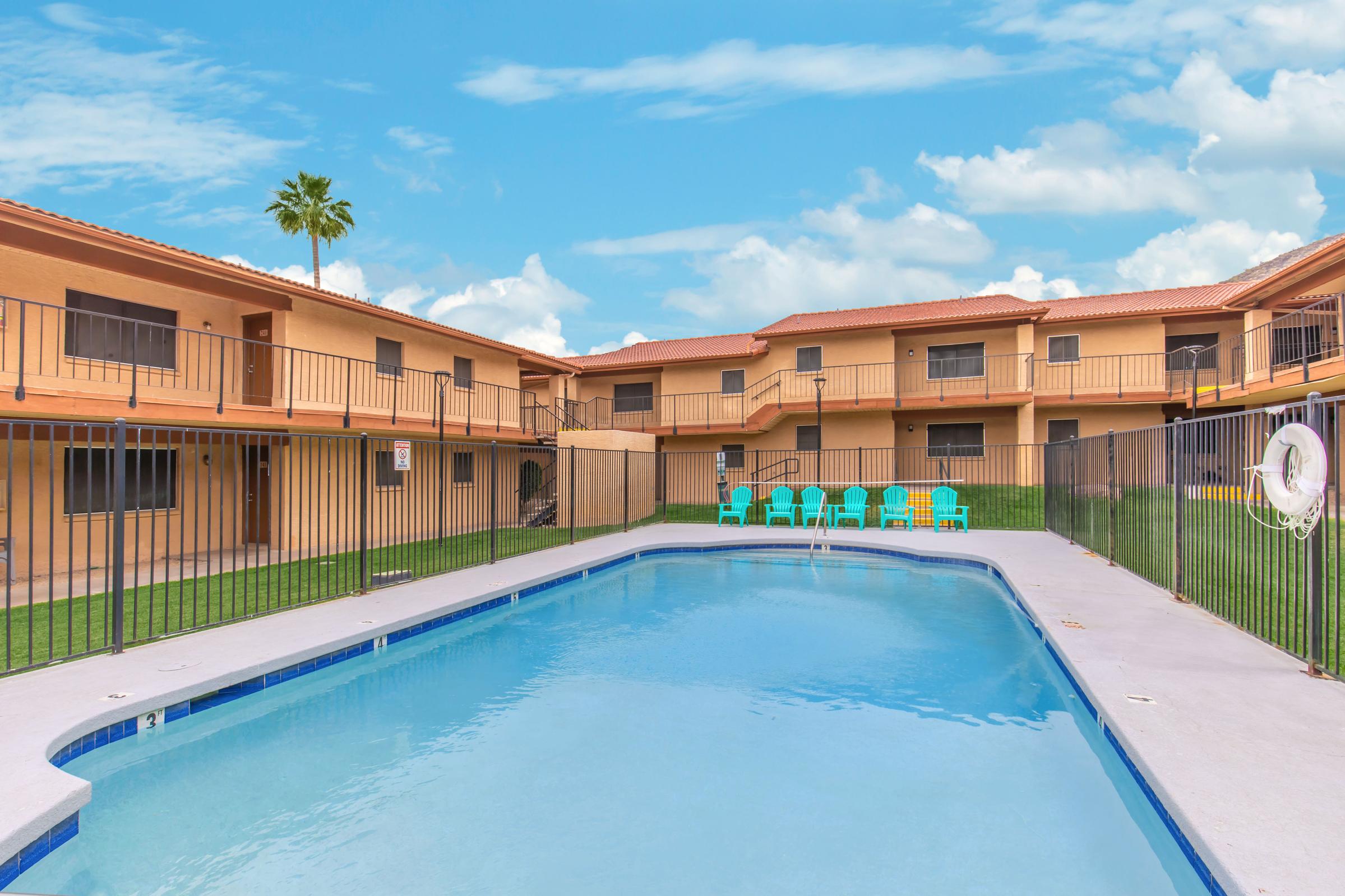 A sunny courtyard featuring a swimming pool surrounded by a fence, with a few lounge chairs nearby. In the background, there are two-story apartment buildings with balconies. A palm tree is visible, set against a blue sky with fluffy clouds.