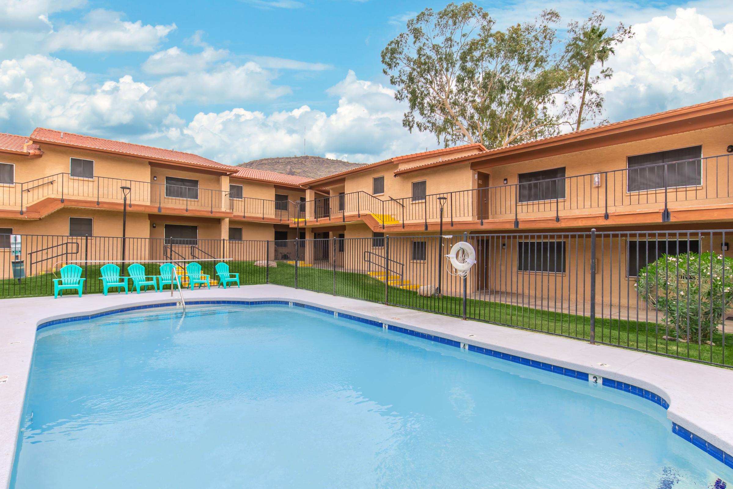 View of a swimming pool surrounded by a well-maintained courtyard featuring green grass and lounge chairs. The backdrop includes two-story apartment buildings with balconies and bright exterior colors under a blue sky with scattered clouds.