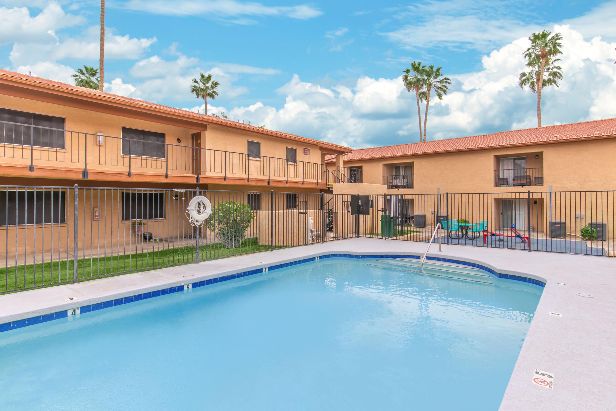 View of a swimming pool surrounded by a courtyard with two-story buildings. The pool is fenced and features a ladder, while colorful lounge chairs are placed nearby. Palm trees are visible in the background under a partly cloudy sky.