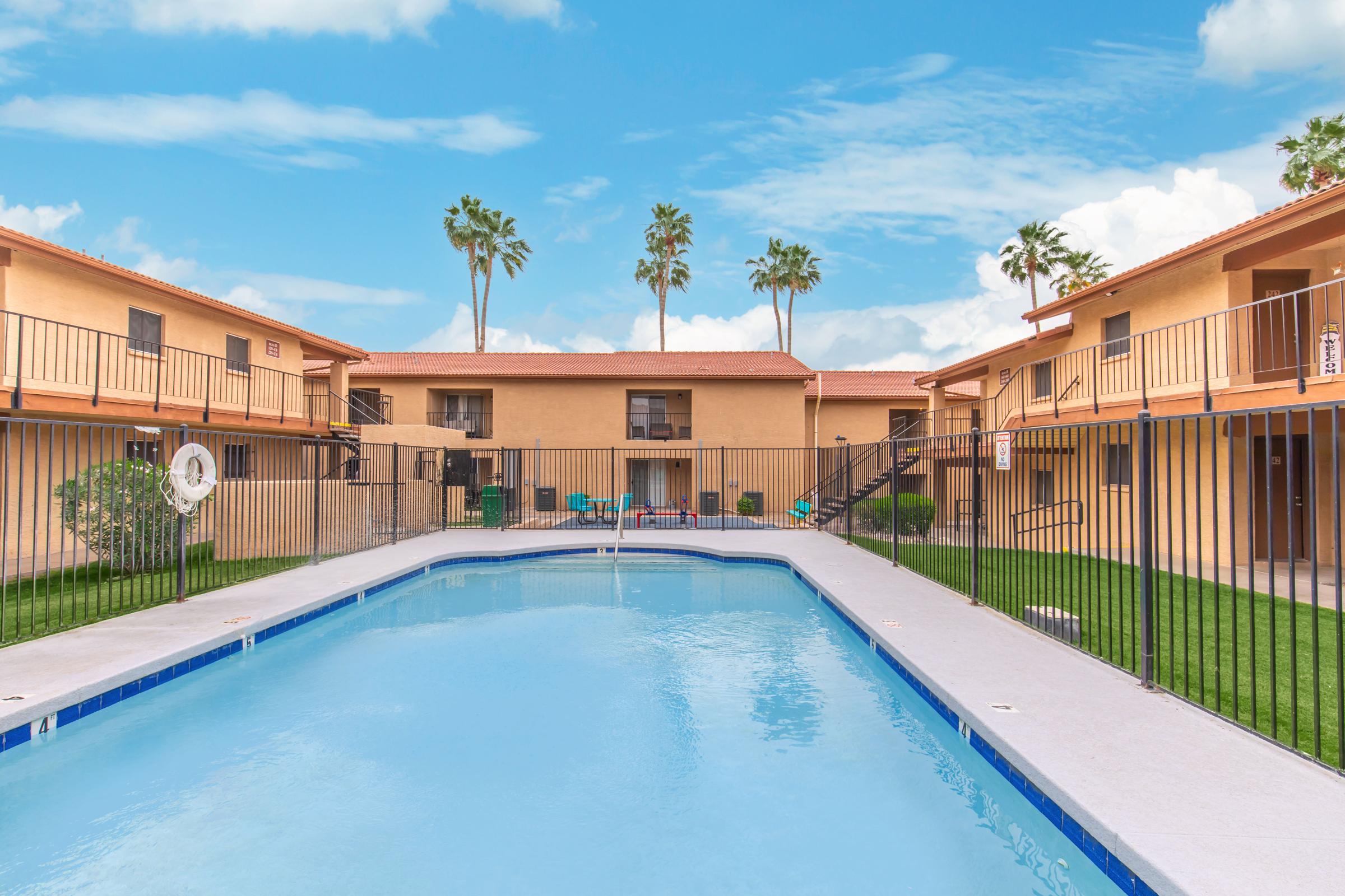 A clear blue swimming pool surrounded by a fenced area, with two-story apartment buildings on either side. Palm trees are visible against a bright blue sky, creating a sunny and inviting atmosphere in a residential complex. Green grass and lounge chairs enhance the poolside setting.