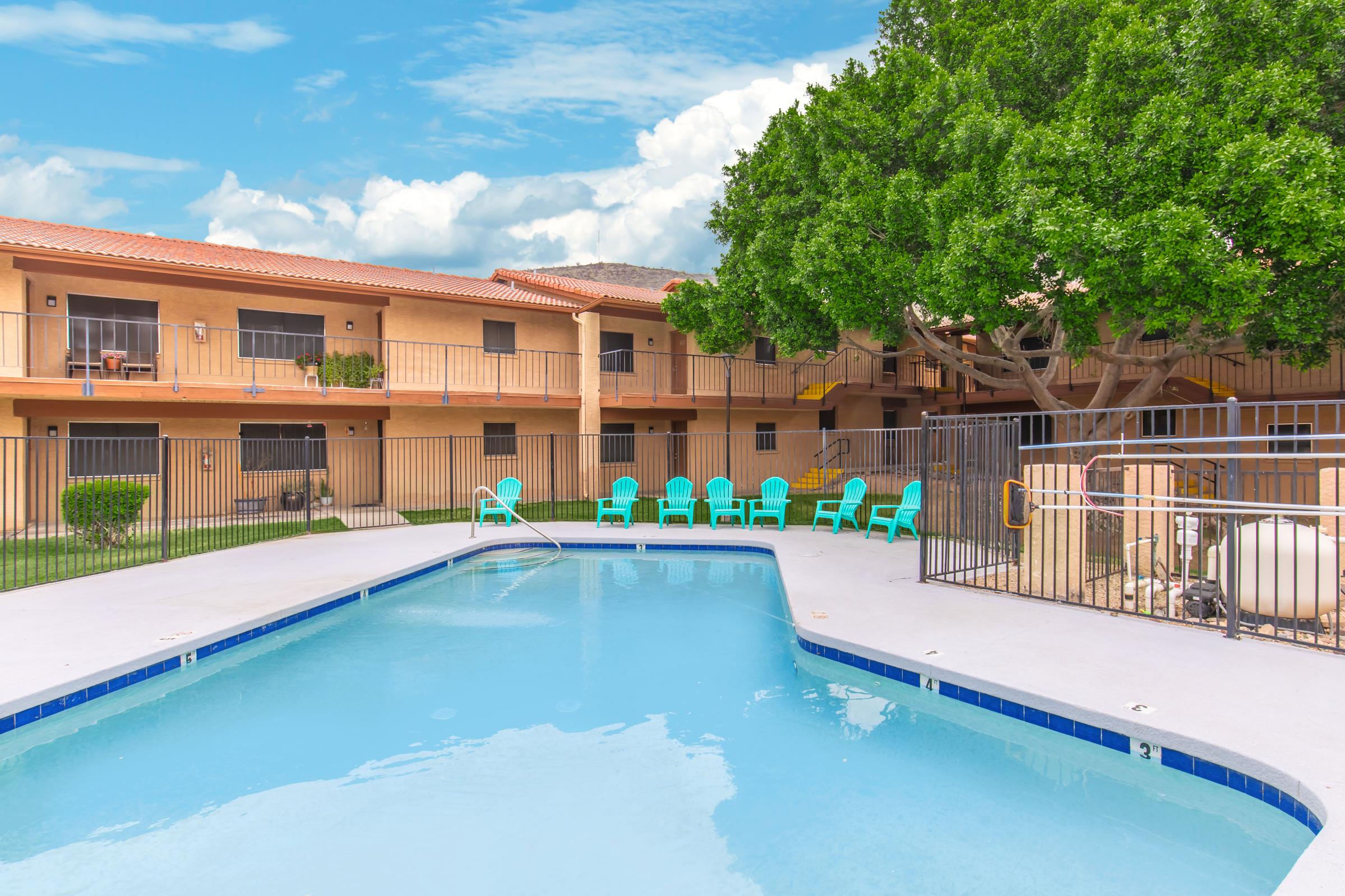 Swimming pool surrounded by colorful adirondack chairs, with a landscaped courtyard featuring a tree. Two-story apartment buildings with balconies are visible in the background against a blue sky with clouds. A fence encloses the pool area, creating a relaxing outdoor space.