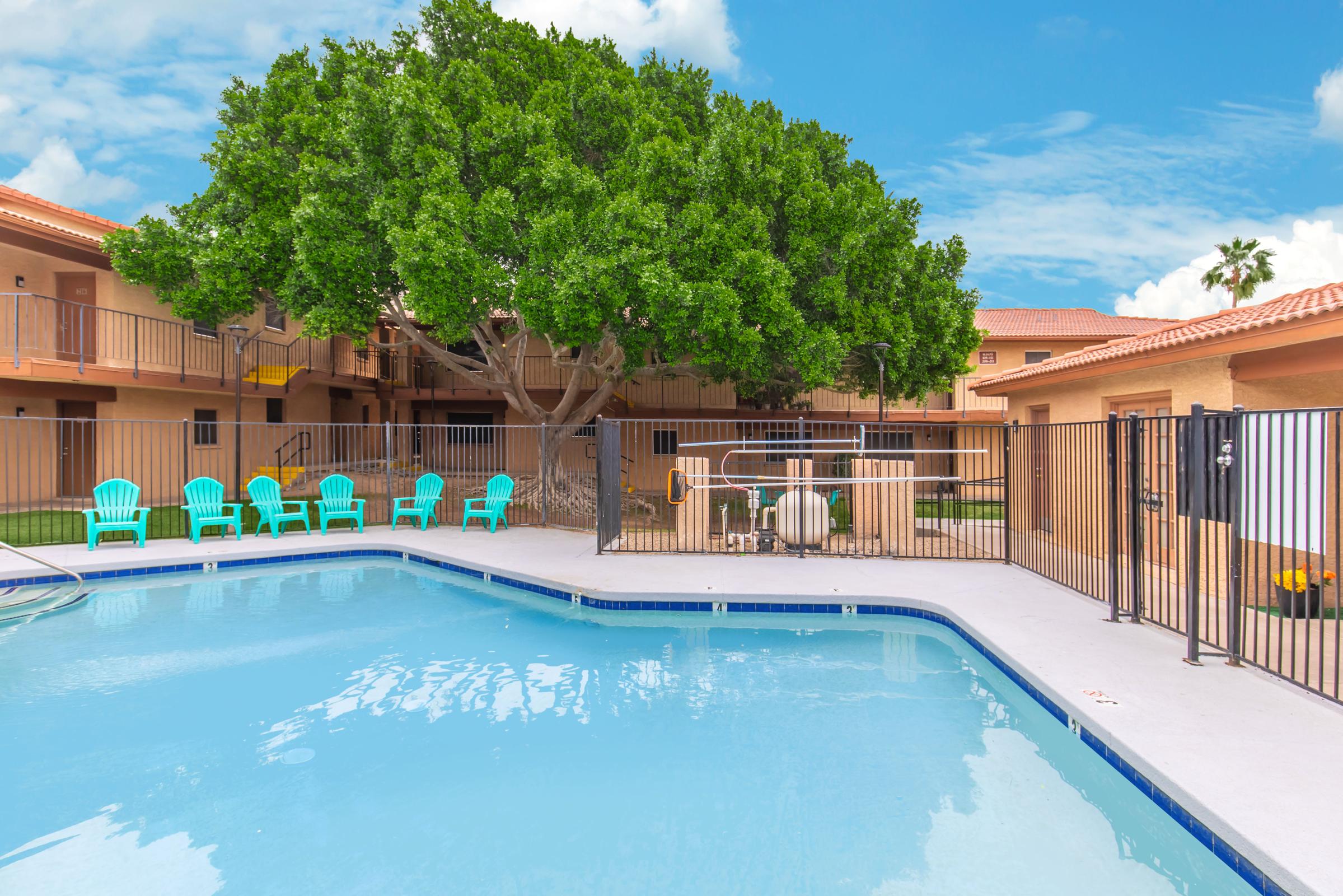 A clear swimming pool surrounded by a fence, with several blue lounge chairs positioned nearby. In the background, there are apartments with tan walls and a large green tree providing shade. The sky is bright blue with fluffy white clouds.