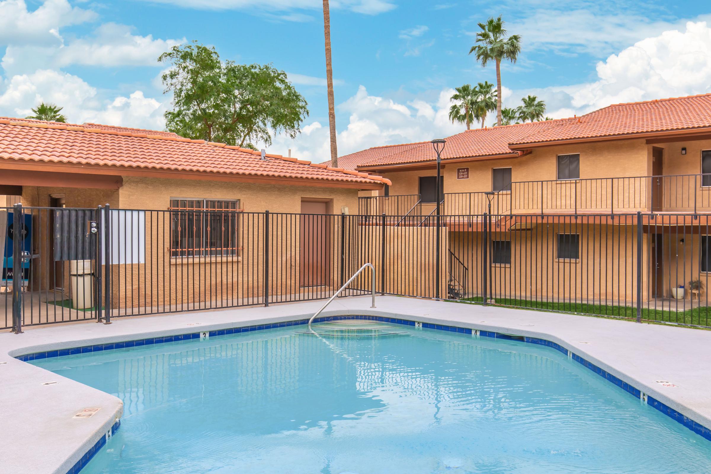 A sunny courtyard featuring a clear blue swimming pool surrounded by orange-walled buildings with tile roofs. The area is enclosed by a black metal fence, and palm trees are visible in the background against a bright blue sky with fluffy white clouds.