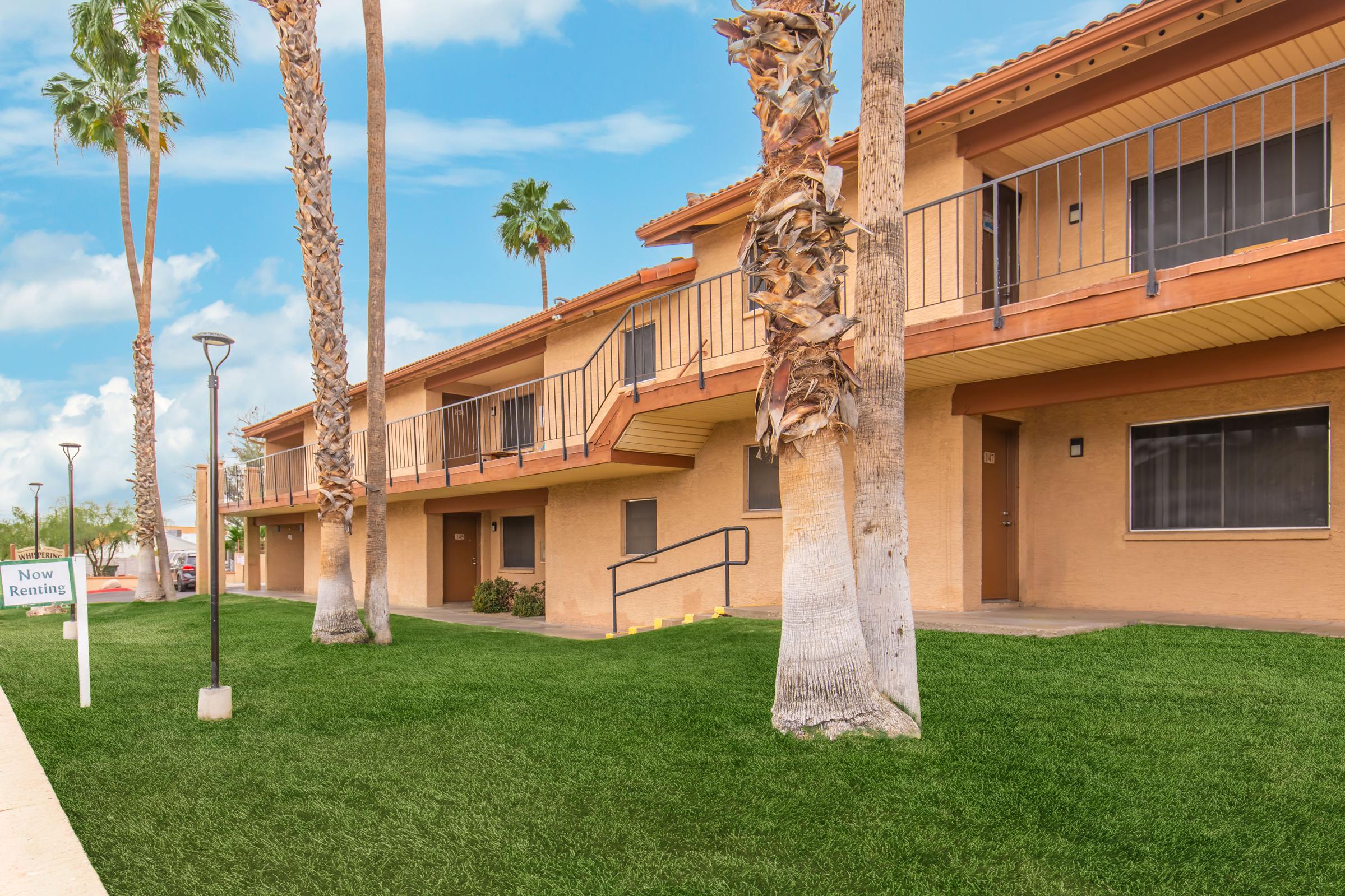 A view of a two-story apartment building with palm trees and a manicured lawn. The building features balconies, and a sign indicating "Now Renting" is visible in front. The sky is clear and blue, contributing to a sunny atmosphere.