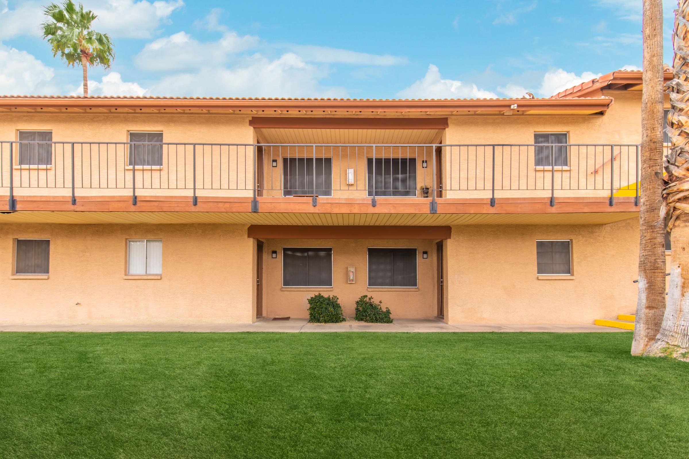 A two-story apartment building with a light brown exterior. It features balconies with black railings, large windows, and grassy areas in front. Palm trees are visible on the sides, and the sky is clear with a few clouds. The entrance doors are centered on the ground floor.