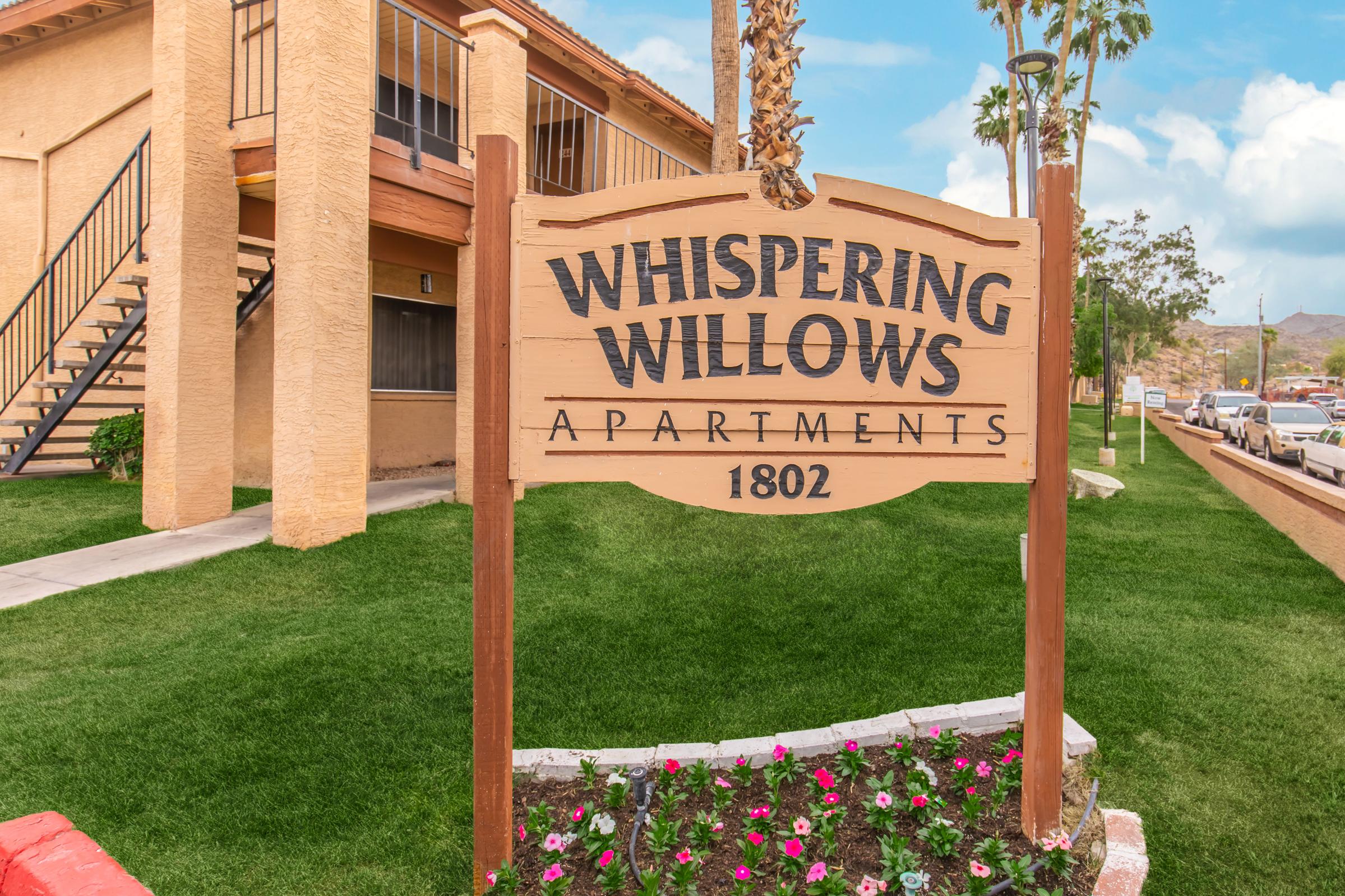 Sign for Whispering Willows Apartments, featuring the name prominently displayed in large letters. The sign is set against a backdrop of a well-maintained grassy area with flowerbeds and a two-story apartment building in the background. Palm trees are visible nearby under a clear blue sky.