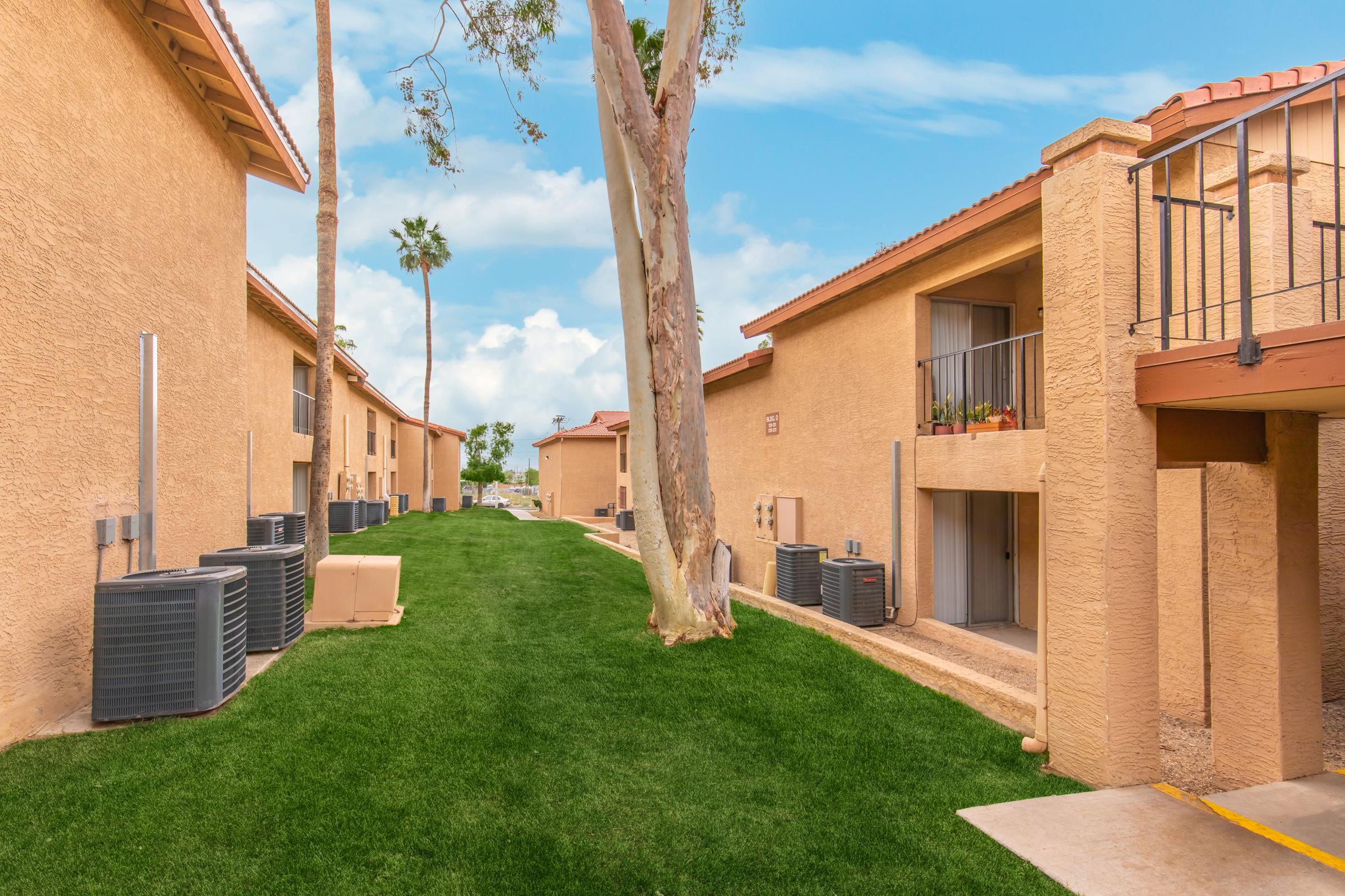 View of a residential area featuring several beige apartment buildings with balconies and green lawns. Air conditioning units are visible along the sides of the buildings, and there are palm trees in the background under a blue sky with fluffy clouds. A peaceful and well-maintained environment.
