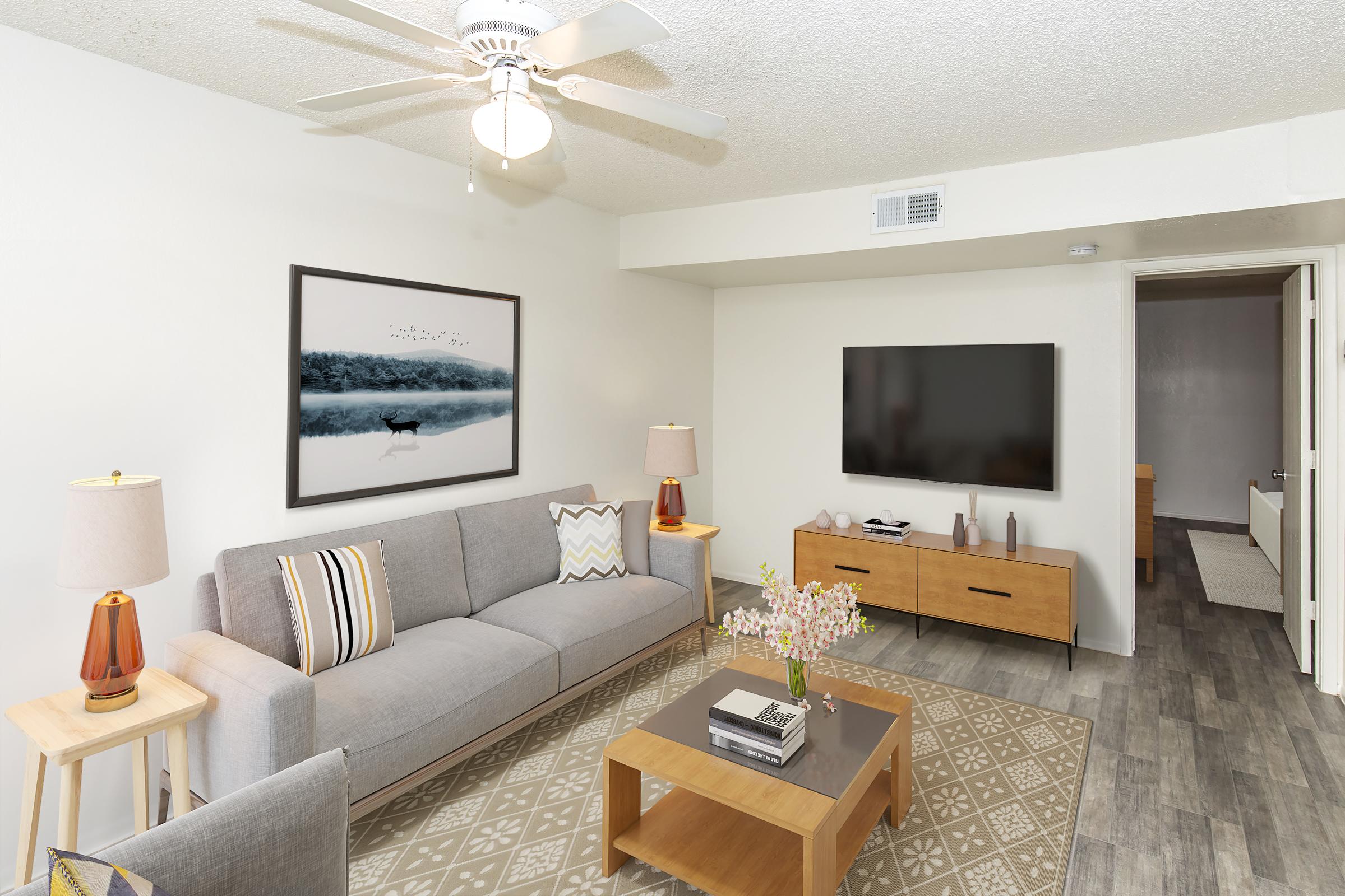A cozy living room featuring a light gray sofa with decorative pillows, a wooden coffee table with a floral arrangement, and a large wall-mounted TV. There are two wooden side tables with lamps, and a framed landscape picture hangs on the wall. The flooring is light wood, and there's a doorway leading to another room.