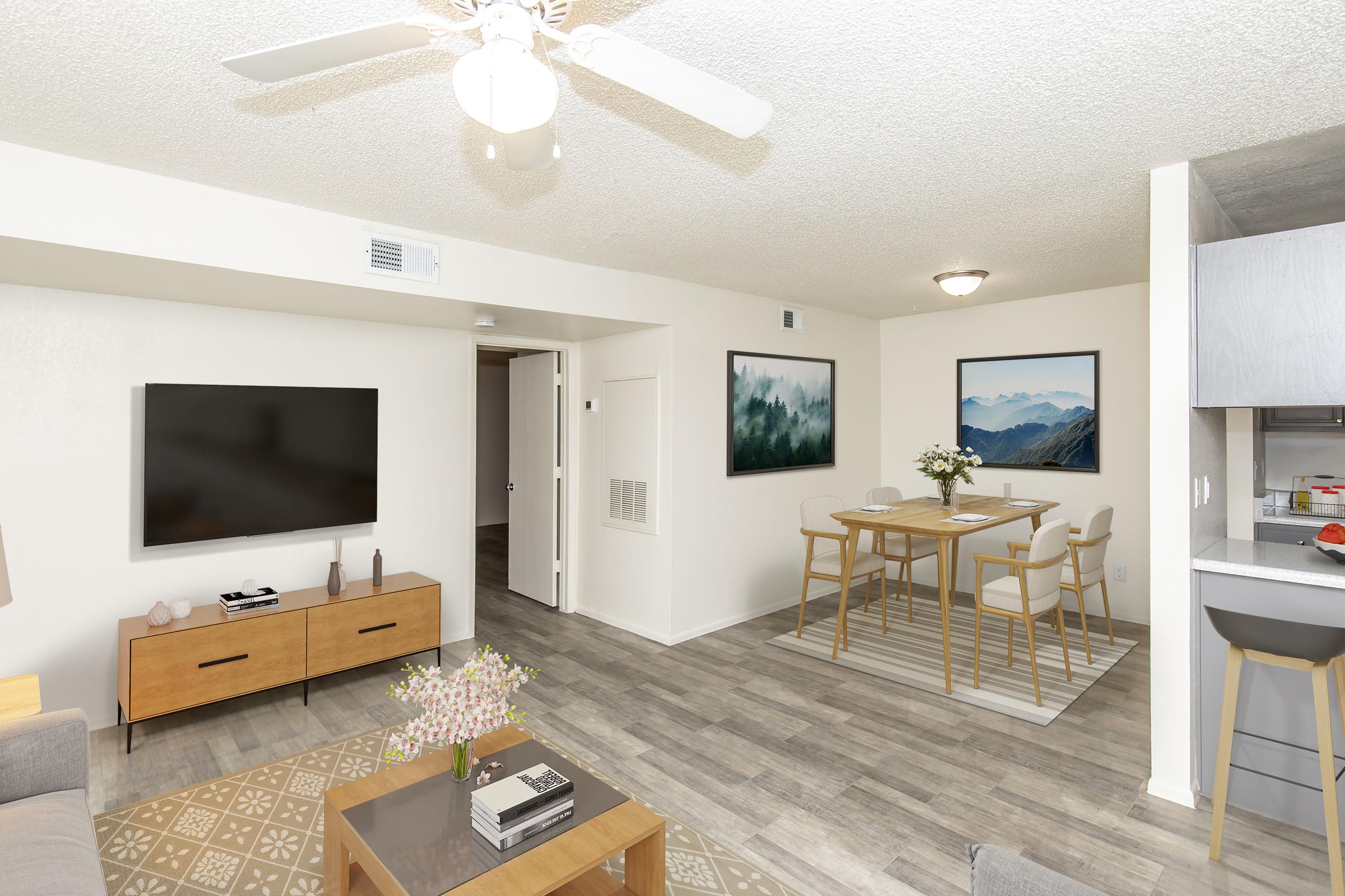 A modern living room and dining area featuring a wall-mounted TV, a wooden coffee table with decorations, and a comfortable couch. The dining space includes a wooden table and chairs. Light-colored walls and large windows create an inviting atmosphere, with artwork and a carpet adding warmth to the interior design.