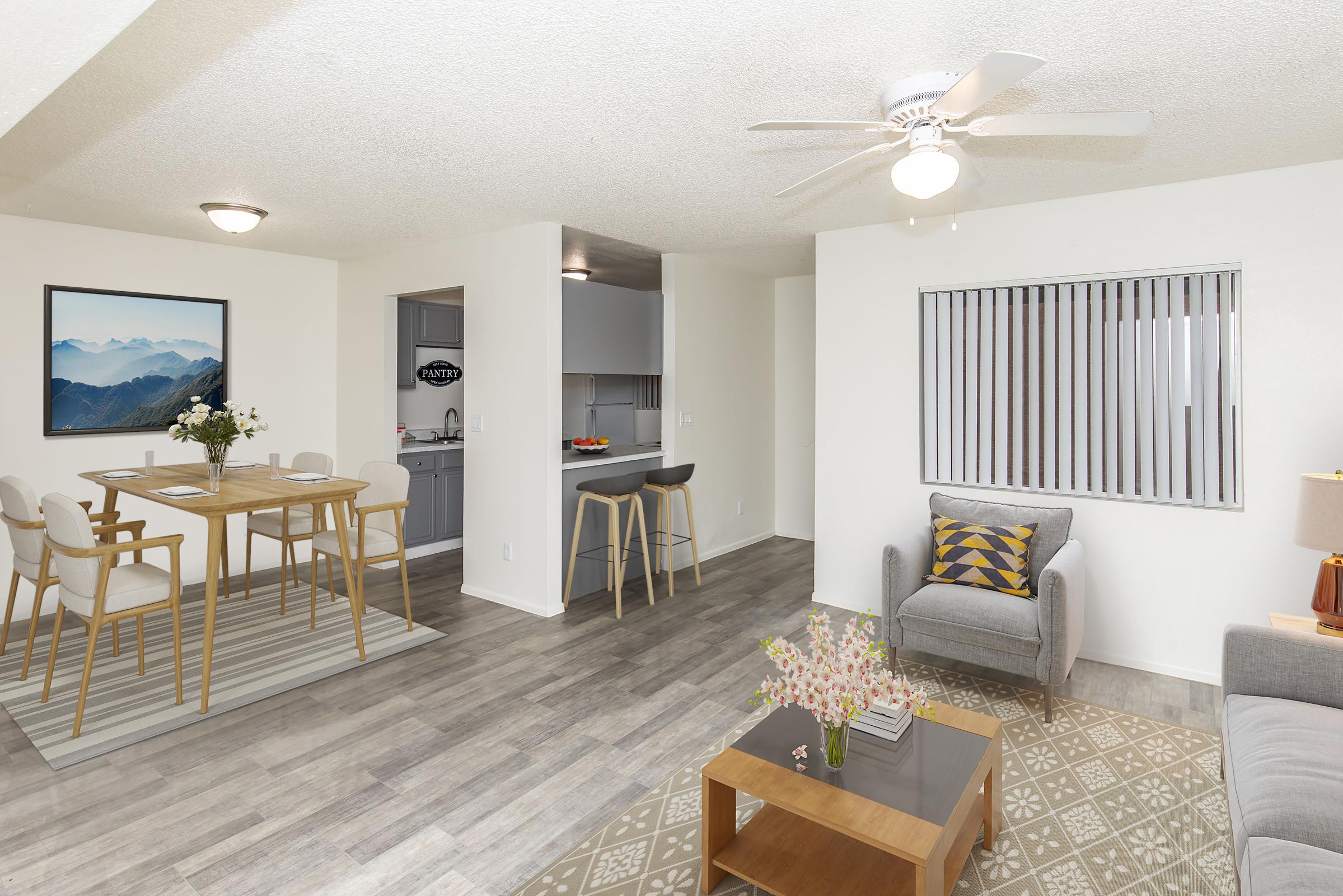 Bright and airy living space featuring a cozy seating area with a gray couch and a coffee table. A dining area with a wood table and chairs is nearby, with modern decor including a ceiling fan and framed mountain artwork. The kitchen is partially visible, showcasing contemporary design elements.