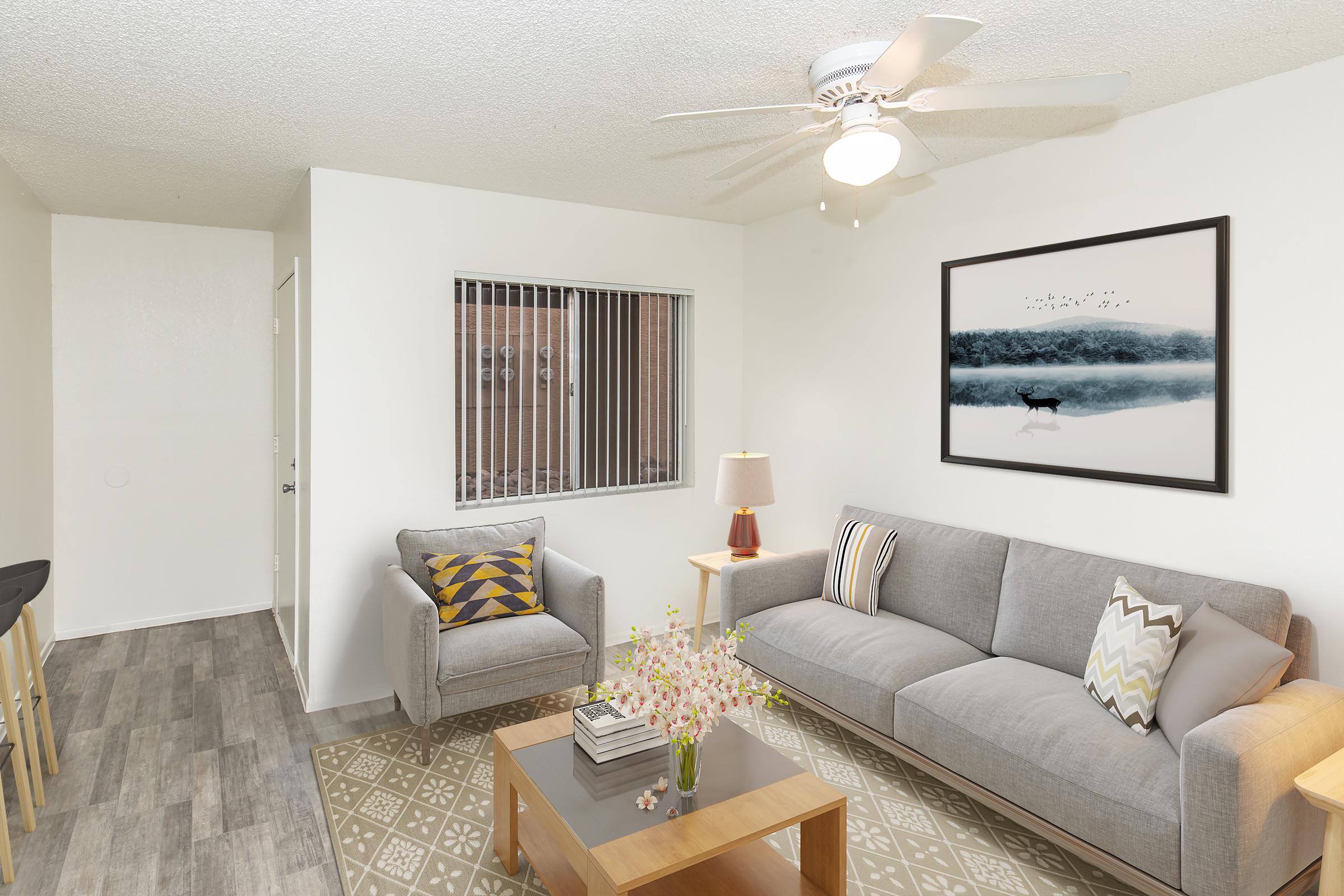 A modern living room featuring a gray sofa with decorative pillows, a small armchair, a wooden coffee table with books and a vase of flowers, and a lamp. A large framed artwork of a serene landscape hangs on the wall. A ceiling fan is visible, and the room has large windows with blinds.