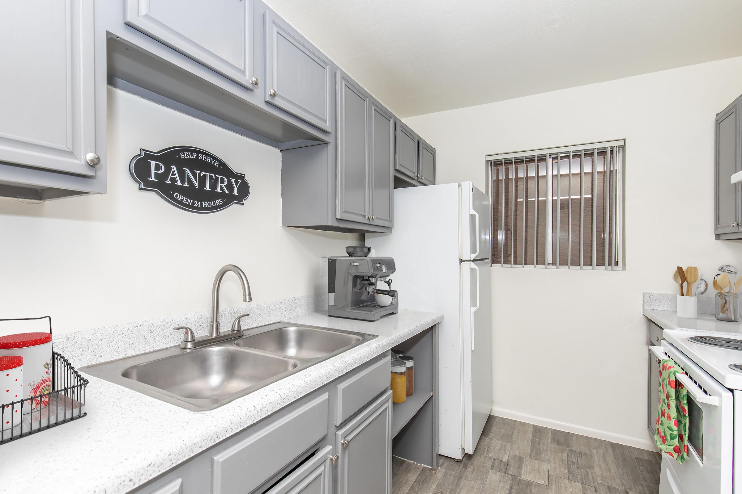 A modern kitchen featuring gray cabinets, a white countertop, and a two-basin sink. There’s a pantry sign on the wall, a white refrigerator, and a coffee maker on the counter. A window with horizontal blinds allows light in, and kitchen utensils and appliances are neatly organized.