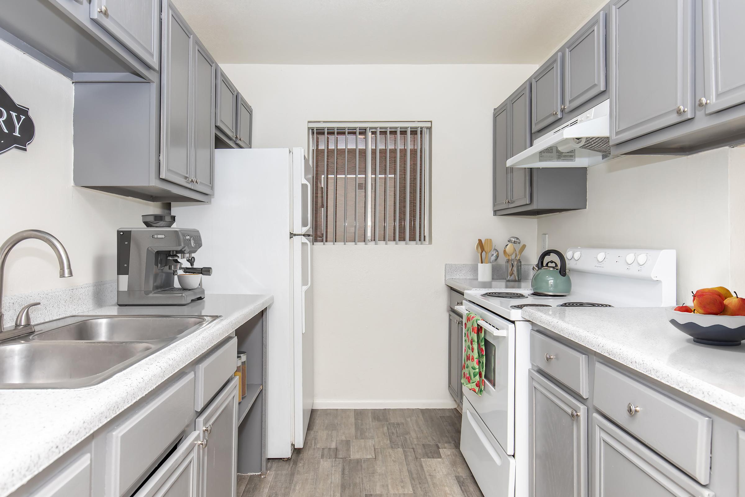 A modern kitchen featuring gray cabinets, a dual sink, a white refrigerator, and a stove with an overhead microwave. A coffee machine sits on the counter, and a green kettle is on the stove. A bowl of apples and kitchen utensils are visible, with a small window providing natural light.