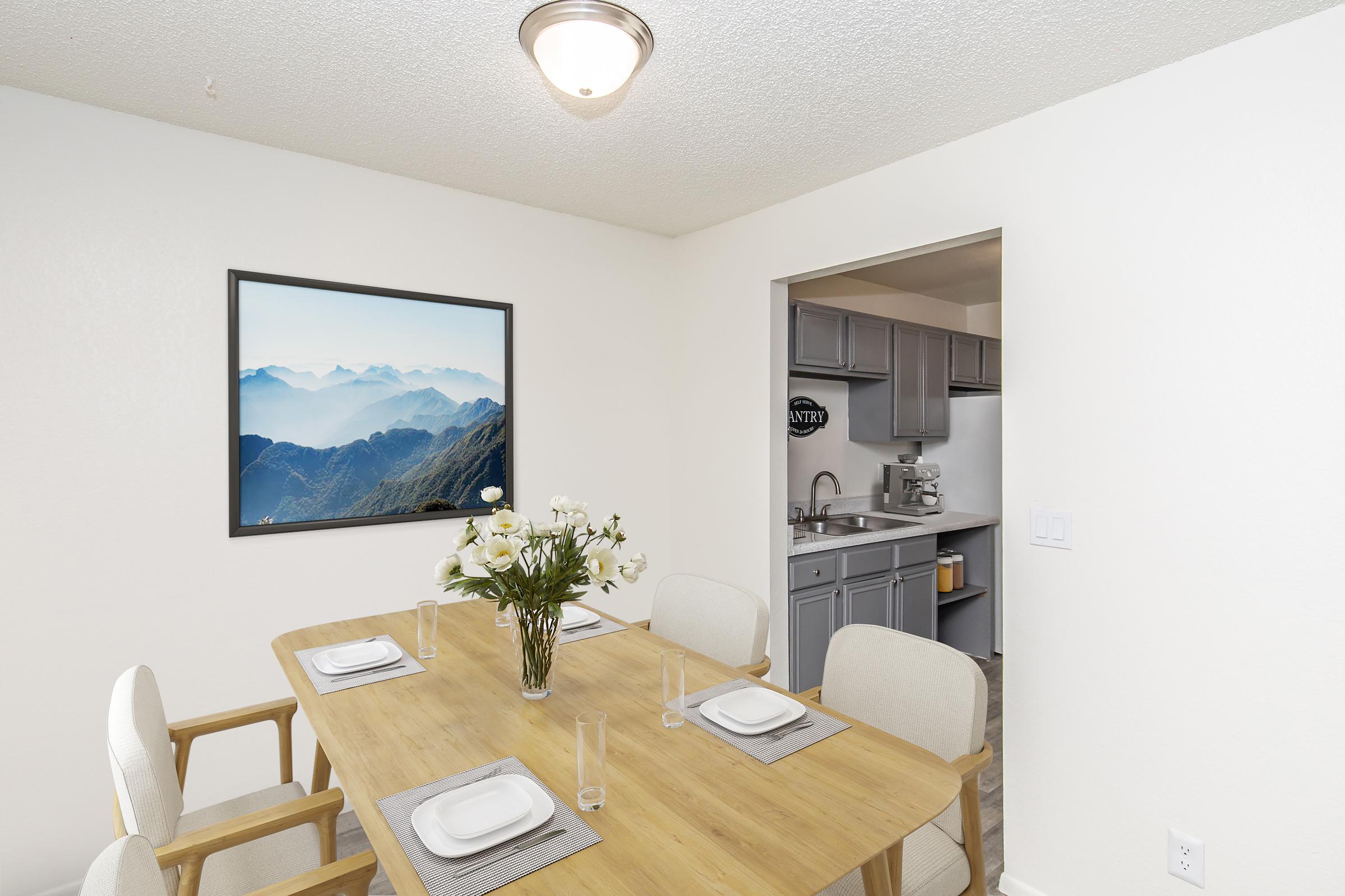 A modern dining area featuring a wooden table set for four with white plates and clear glasses. A vase of white flowers is centerpiece. On the wall, there's a large framed landscape photograph of mountains in a blue haze. A doorway leads to a small kitchen area with gray cabinets and appliances visible.