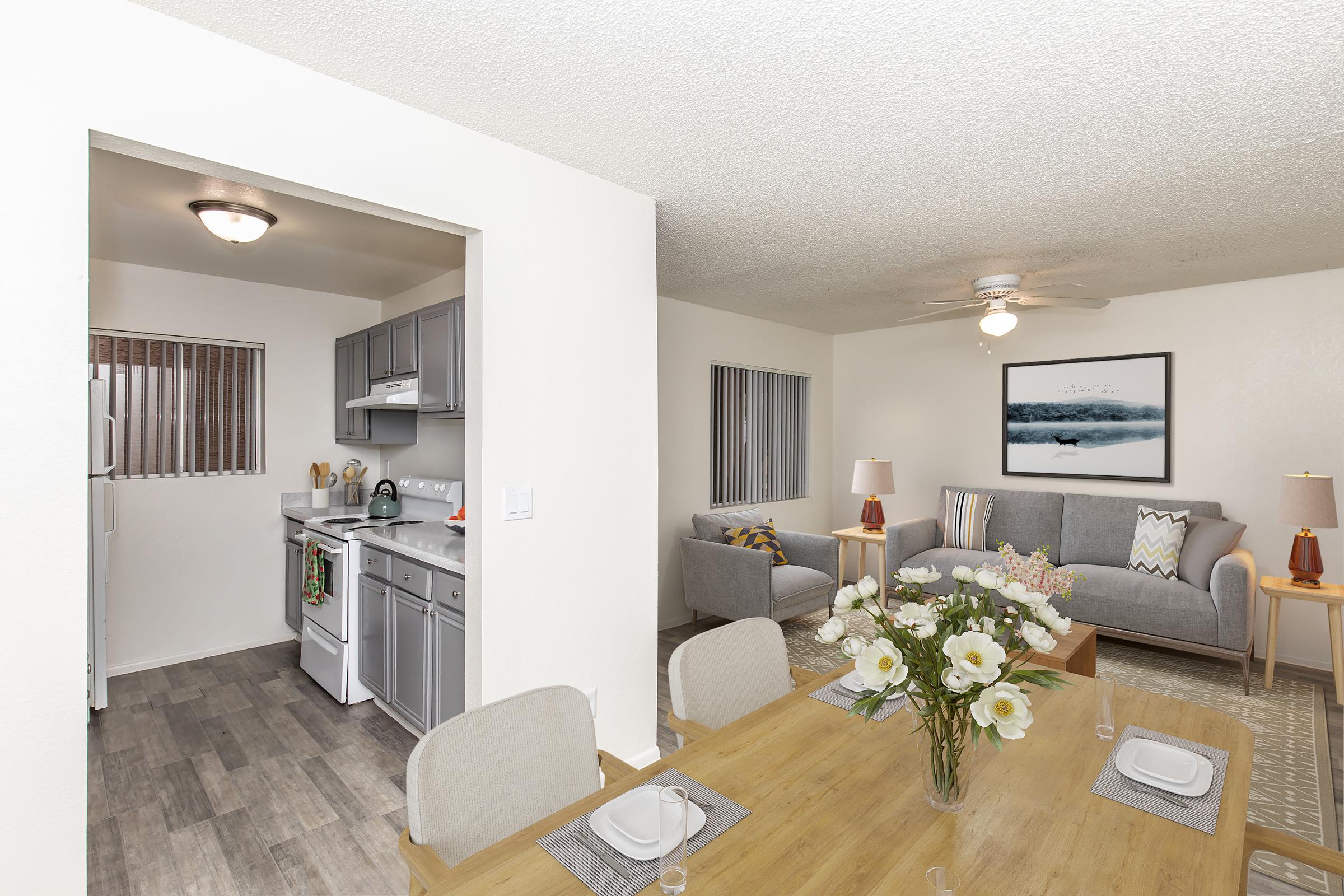 A modern open-concept living space featuring a kitchen with gray cabinets and appliances, a dining table set for four with a vase of flowers, and a cozy living area with a sofa, chairs, and artwork on the wall. Natural light enters from windows, enhancing the inviting atmosphere.