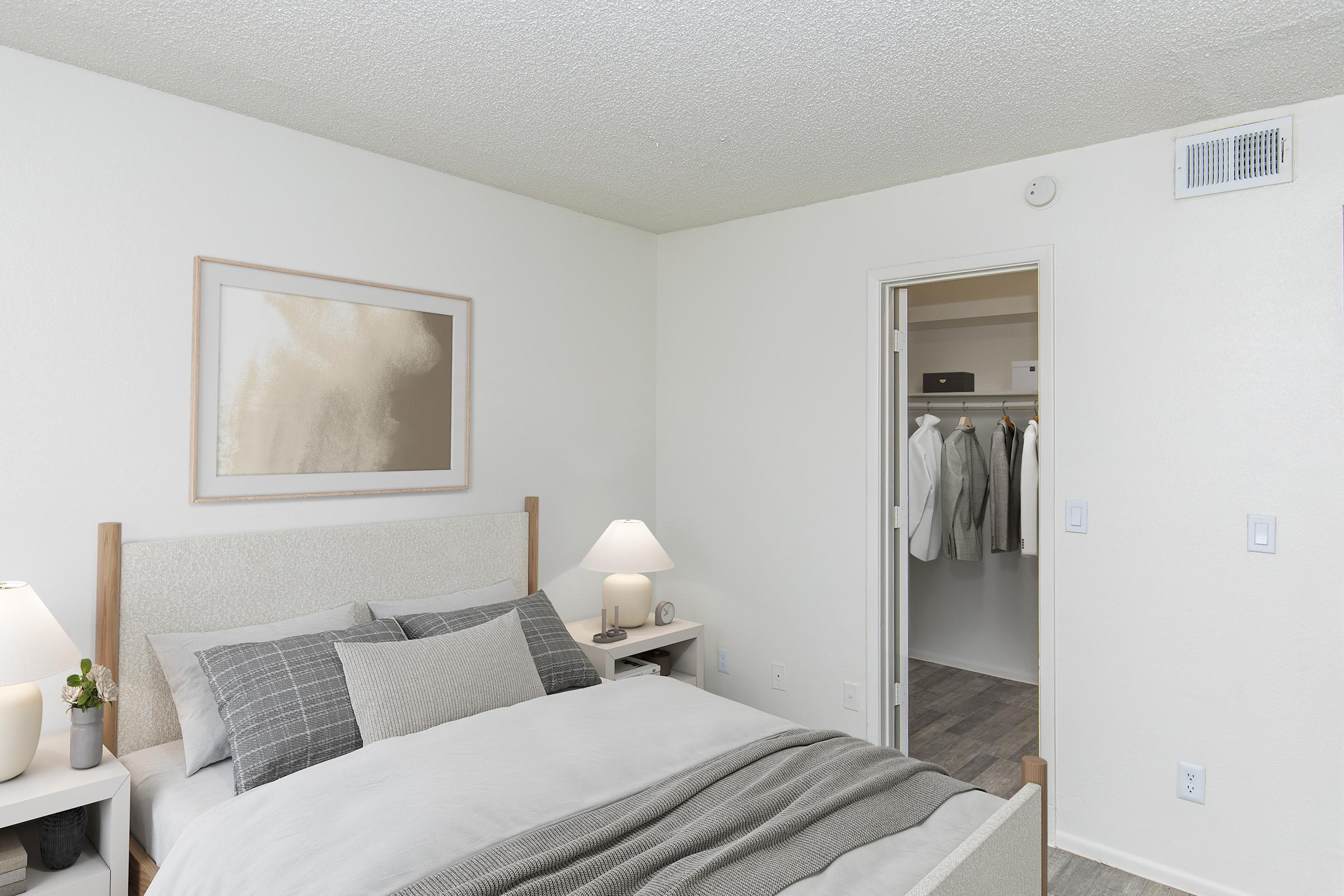 A cozy bedroom featuring a neatly made bed with gray and white bedding, two bedside lamps, and a framed artwork on the wall. A closet with neatly hung clothes is visible through an open door. The room has a neutral color palette and a comfortable, inviting atmosphere.