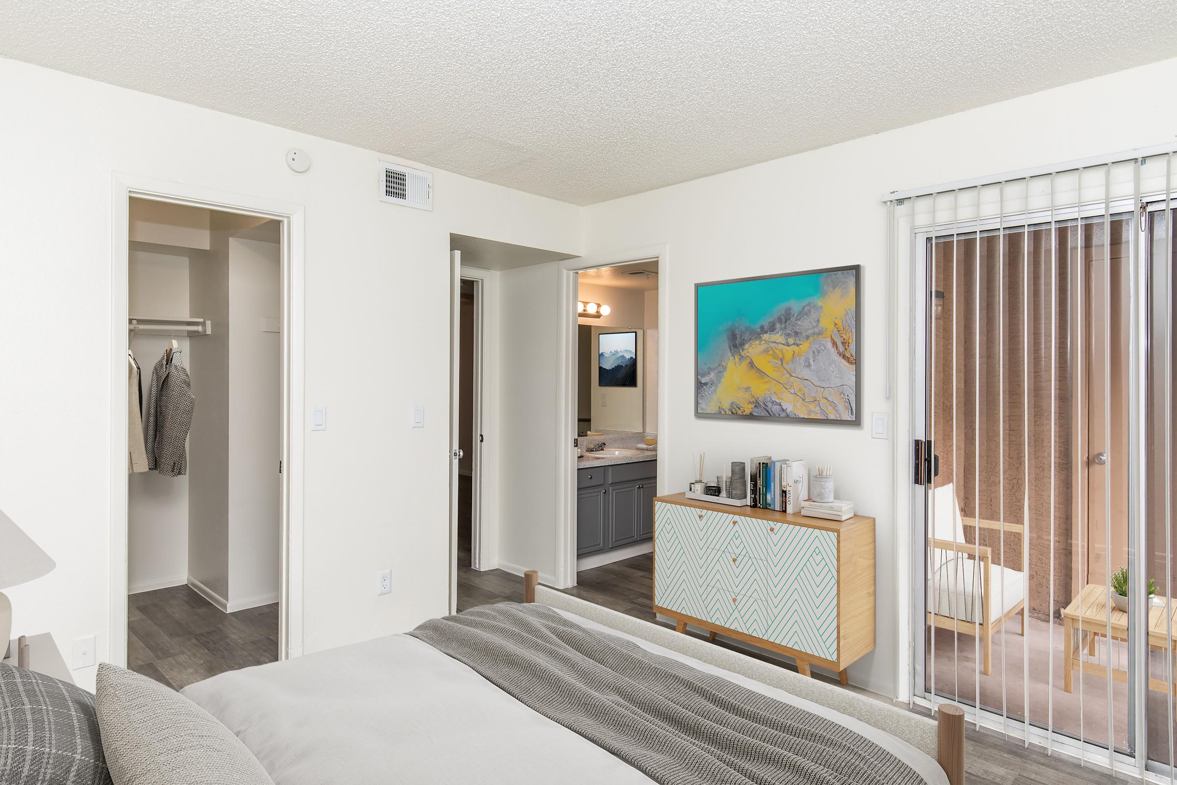 A cozy bedroom featuring a bed with gray bedding, a dresser with decorative items, and an open closet. The room connects to a bathroom area visible through an open door. A sliding glass door leads to an outdoor space, enhancing the natural light in the room. Neutral walls and modern decor create a welcoming atmosphere.