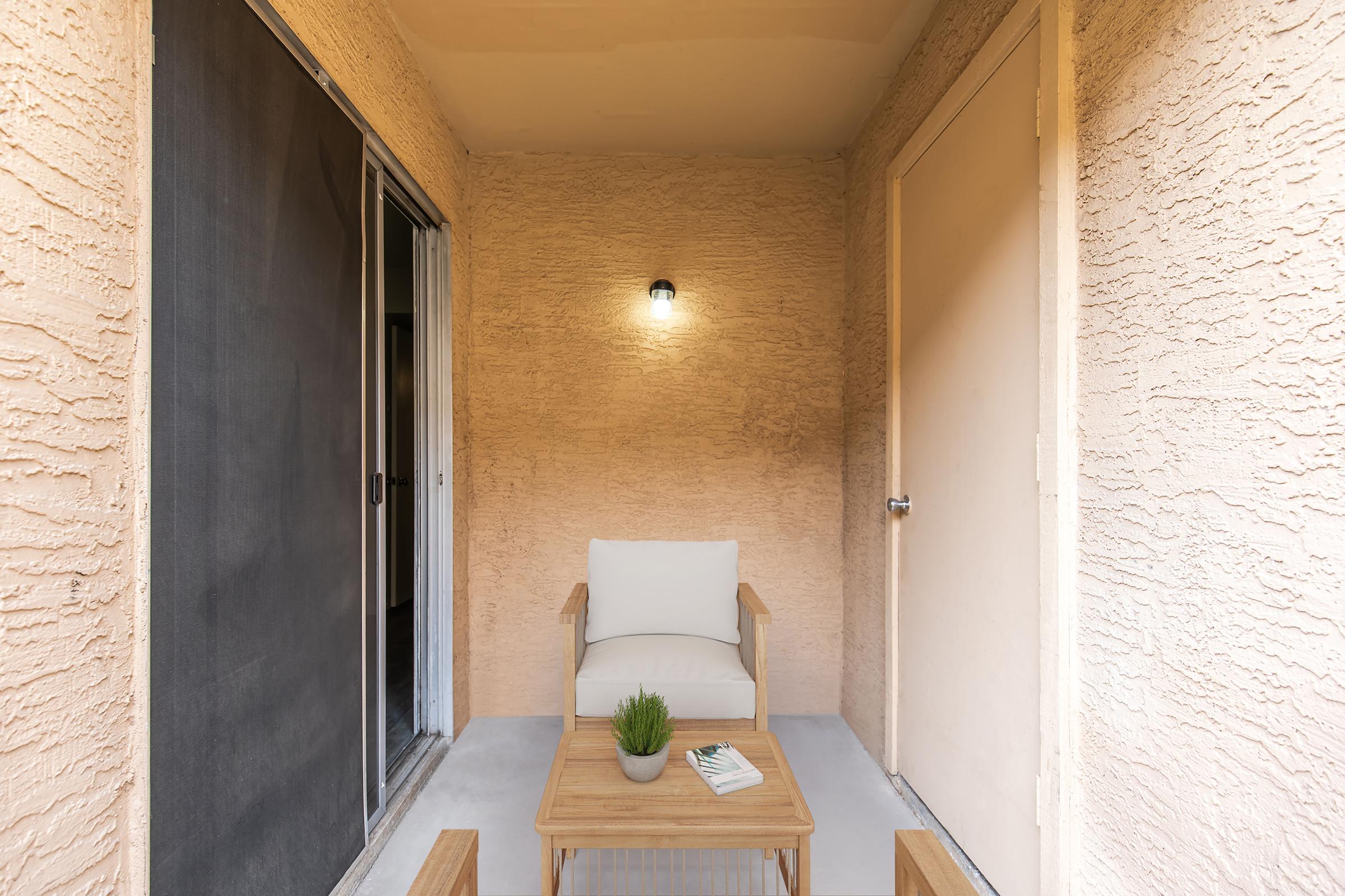 A cozy outdoor patio area featuring a light-colored armchair with a white cushion, a small wooden table with a plant, and a warm-toned textured wall. A sliding door leads to the interior space, and a wall-mounted light fixture provides illumination. The overall ambiance is inviting and relaxed.