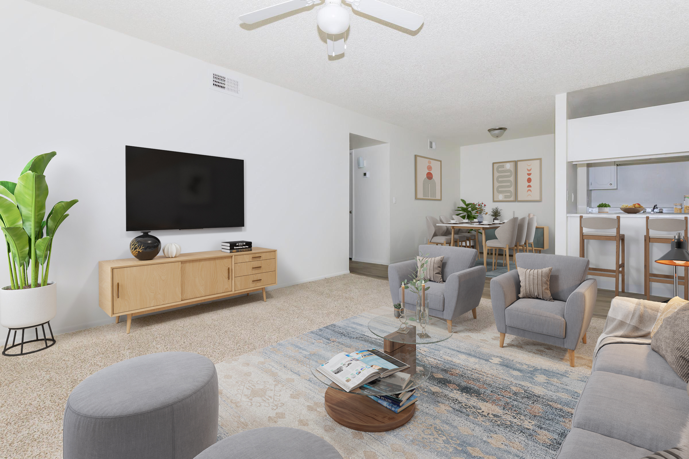 A modern living room featuring a large flat-screen TV mounted on the wall, a beige sofa set with two armchairs, a glass coffee table, and a decorative rug. A potted plant adds greenery, and a dining area with a wooden table and chairs is visible in the background. The space is well-lit and airy.