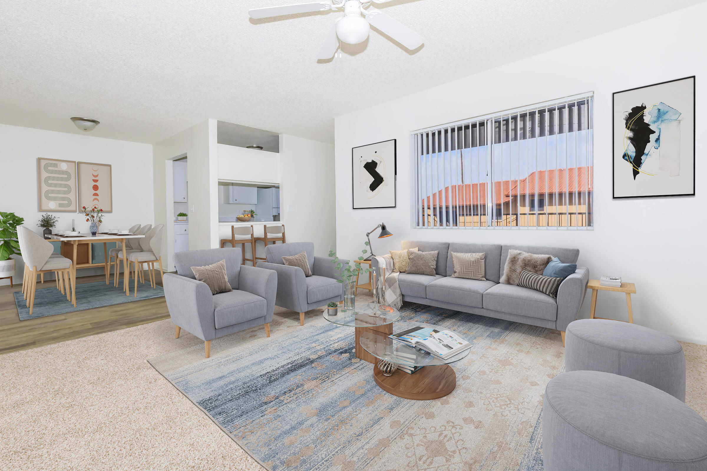 A modern living room featuring a gray sectional sofa with decorative cushions, a round coffee table, and two gray ottomans. Light-colored walls and a large window allow natural light. The décor includes framed art on the walls, a rug beneath the furniture, and a dining area visible in the background.