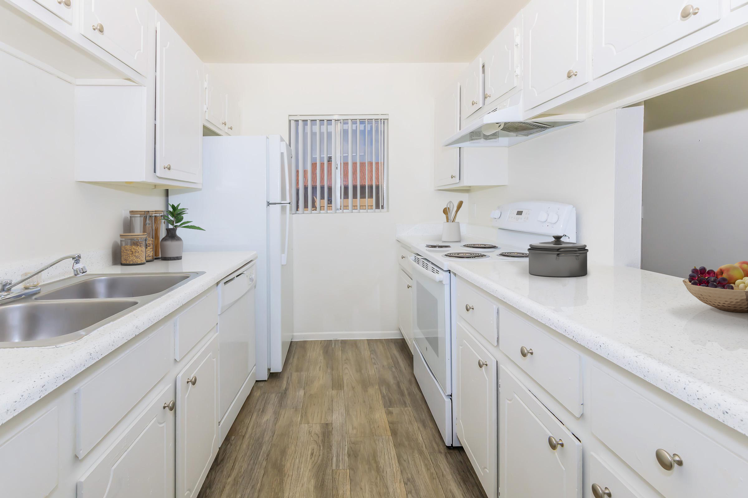 A clean and modern kitchen featuring white cabinetry, a double sink, and stainless steel appliances. The countertops are light-colored with a subtle pattern. A window provides natural light, and there are bowls of fruit and kitchen appliances on the countertops. The floor is a warm wood-like laminate.