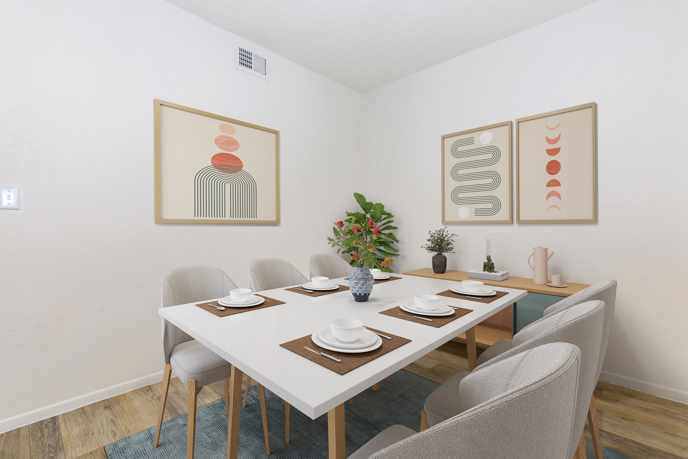 A modern dining room featuring a white table set for six with plates, utensils, and placemats. Two framed abstract art pieces hang on the wall. A potted plant adds greenery, and a vase with flowers sits on the table. The space has light-colored walls and a wooden floor with a blue area rug.