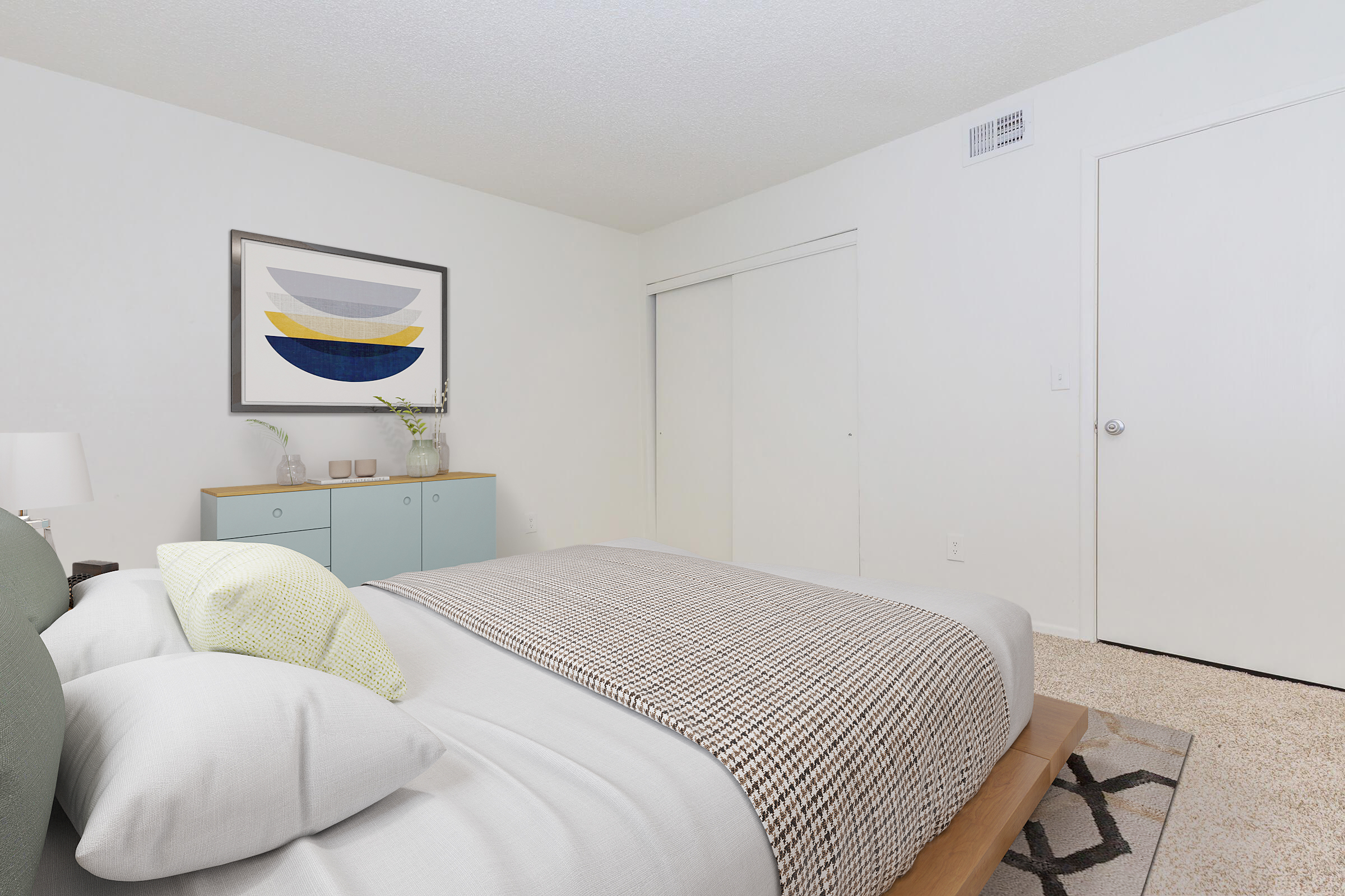 A cozy bedroom featuring a neatly made bed with gray and white bedding, accented by a green pillow. A light blue dresser is positioned against the wall, topped with decorative items. A framed abstract artwork hangs above the dresser, and a closed closet door along with a white door are visible in the background. Soft beige carpet covers the floor.