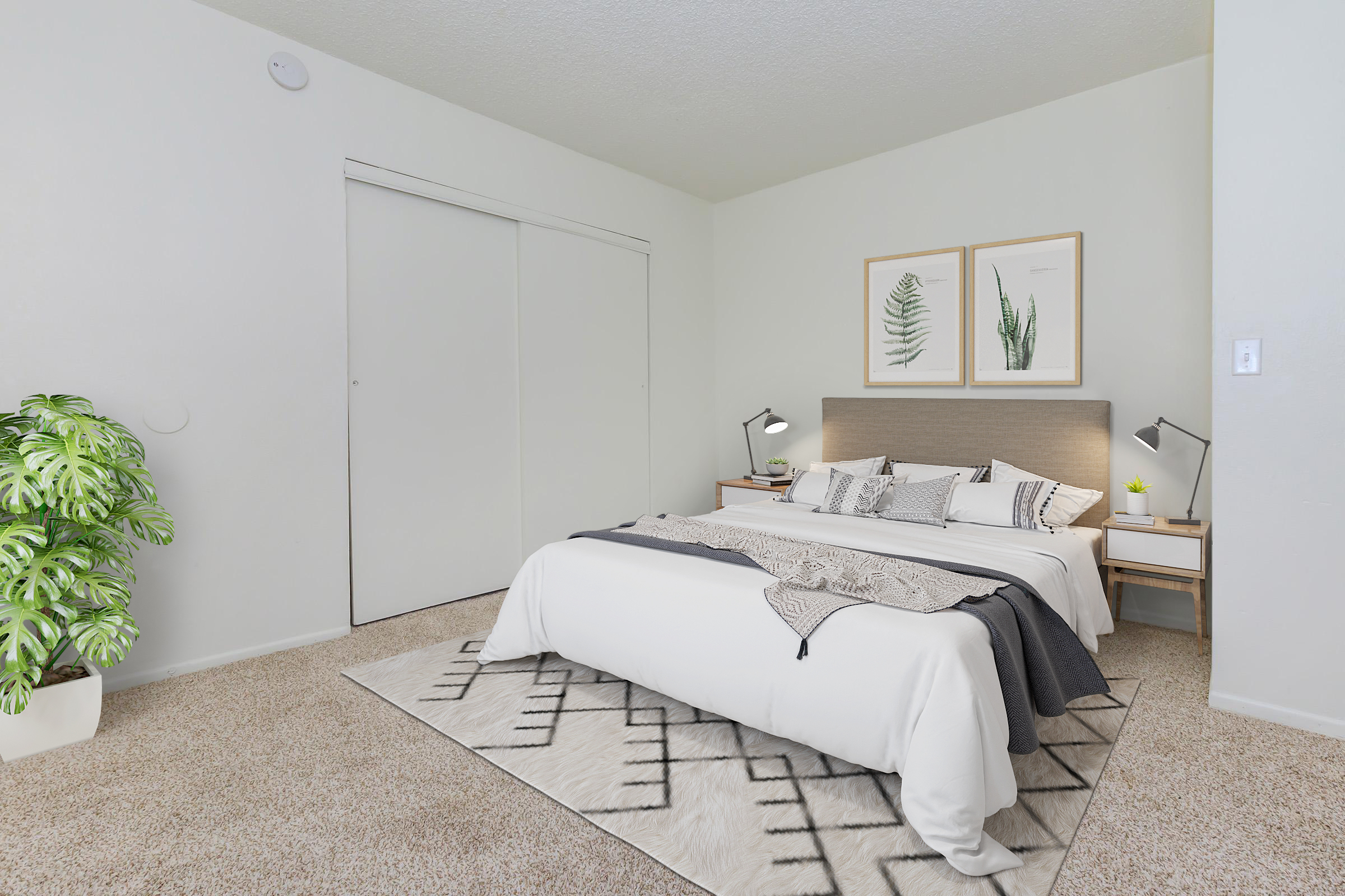 Modern bedroom featuring a king-sized bed with decorative pillows, a stylish headboard, and two framed botanical prints on the wall. A bedside table with lamps sits on either side of the bed, and a large area rug is laid on light carpet. A green plant adds a touch of nature to the space.
