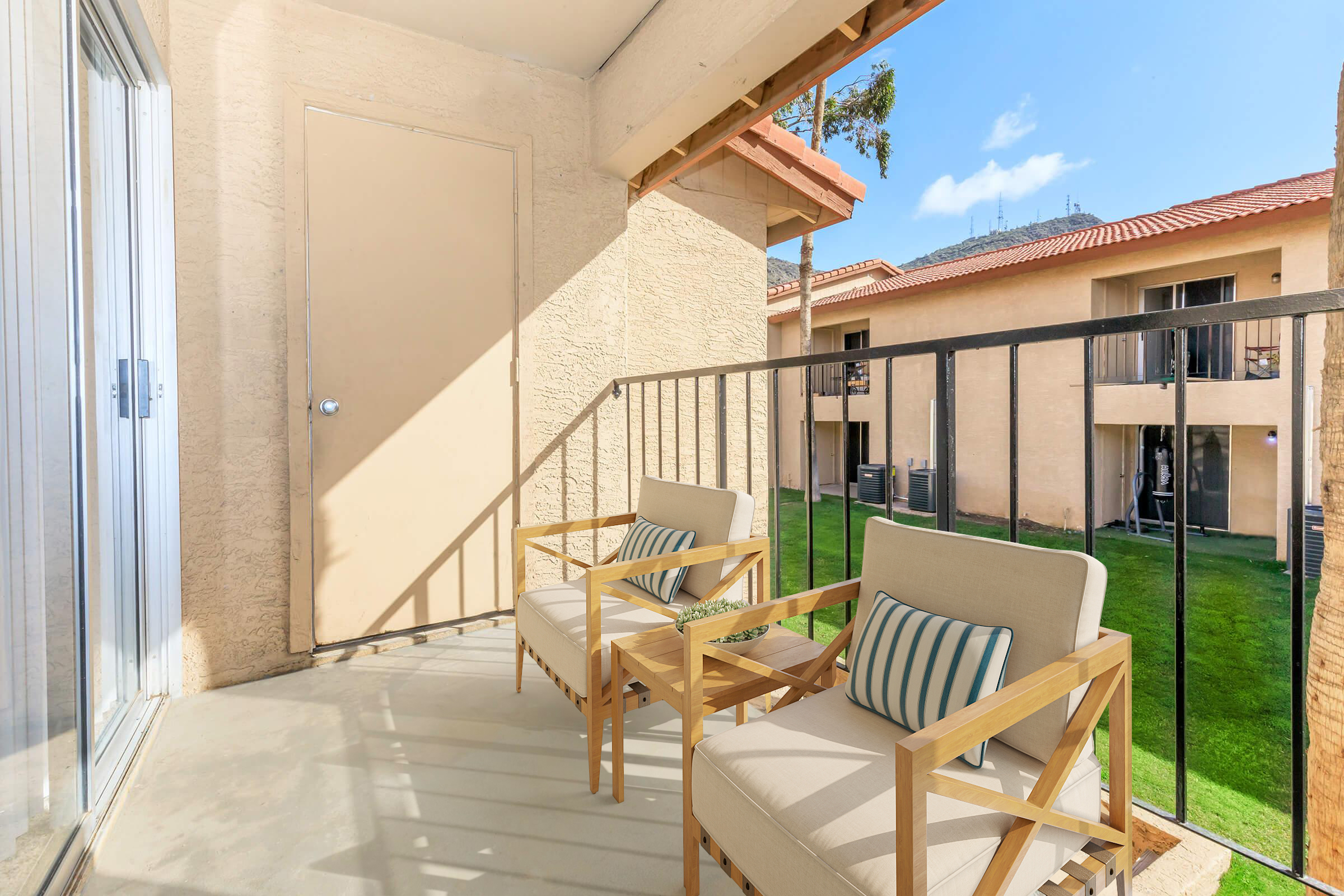 Balcony area featuring two modern chairs with striped cushions, overlooking a grassy courtyard. A beige wall and door are visible, along with a sunny blue sky and adjacent building in the background. The setting suggests a comfortable outdoor space for relaxation.