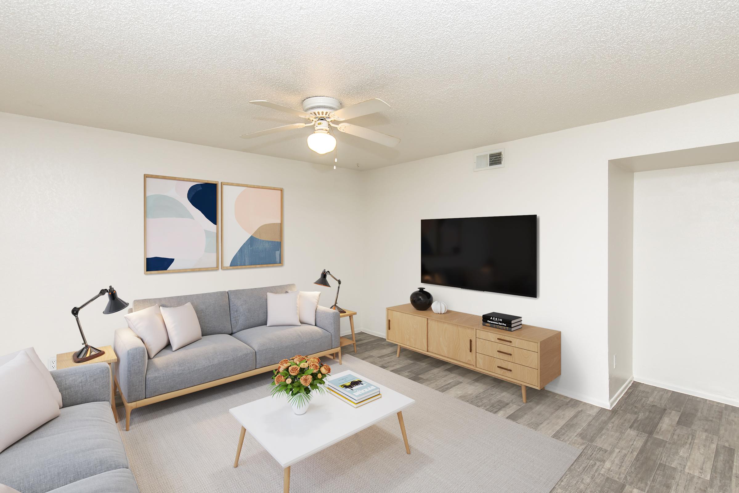 A modern living room featuring a light gray sofa, wooden coffee table with a decorative vase, and two stylish lamps. On the wall are two abstract paintings, and a large flat-screen TV is mounted opposite the seating area. The floor is covered with light-colored tiles, and a ceiling fan adds to the contemporary decor.