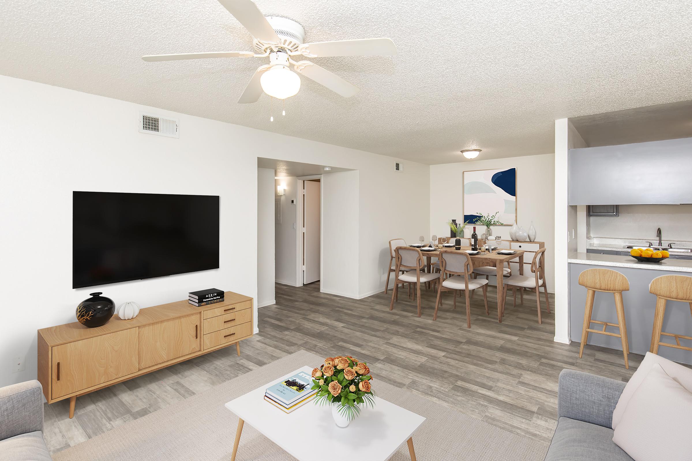 A bright and modern living room featuring a ceiling fan, a large TV mounted on the wall, a stylish coffee table with a vase of flowers, and a comfortable gray sofa. In the background, there is a dining area with a wooden table set for a meal, showcasing an inviting and cozy atmosphere.