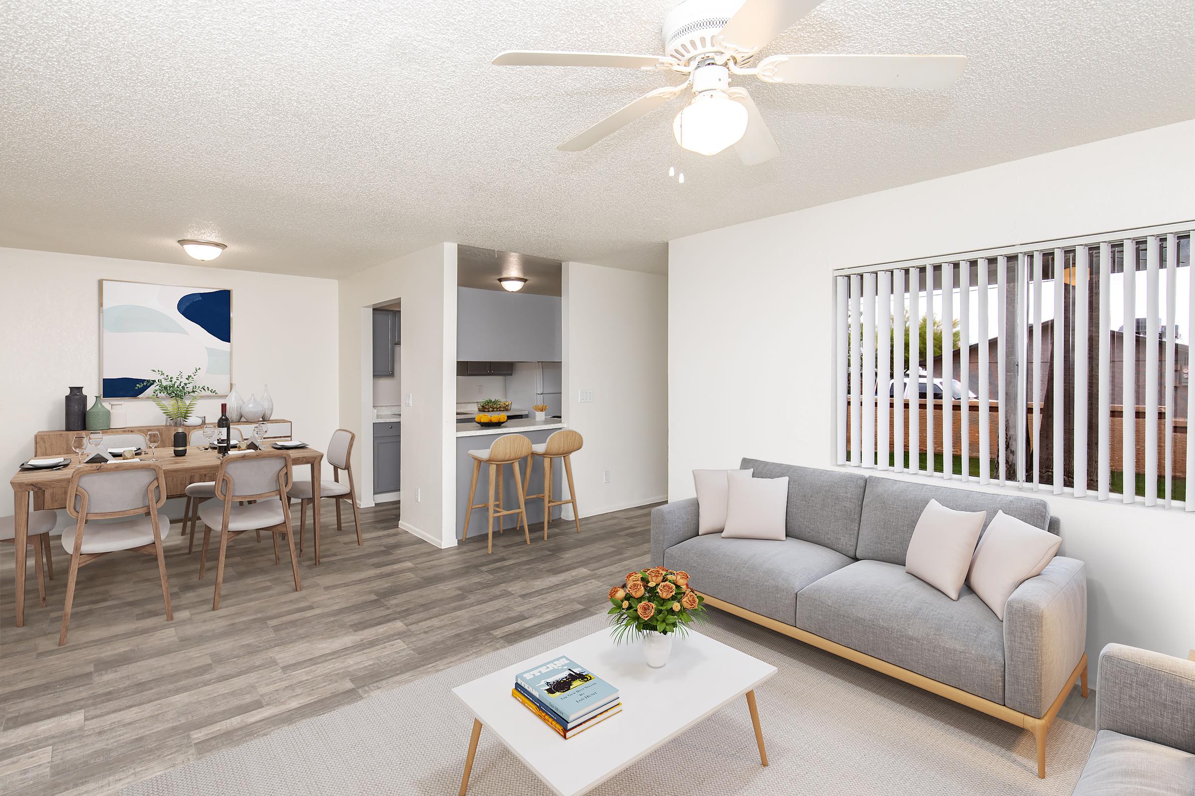 A modern, bright living space featuring a cozy gray sofa with decorative pillows, a coffee table with a plant and books, and a dining area with a wooden table and chairs. A ceiling fan hangs overhead, and sliding blinds cover the window, allowing for natural light. The walls are painted in a light color, enhancing the airy feel.