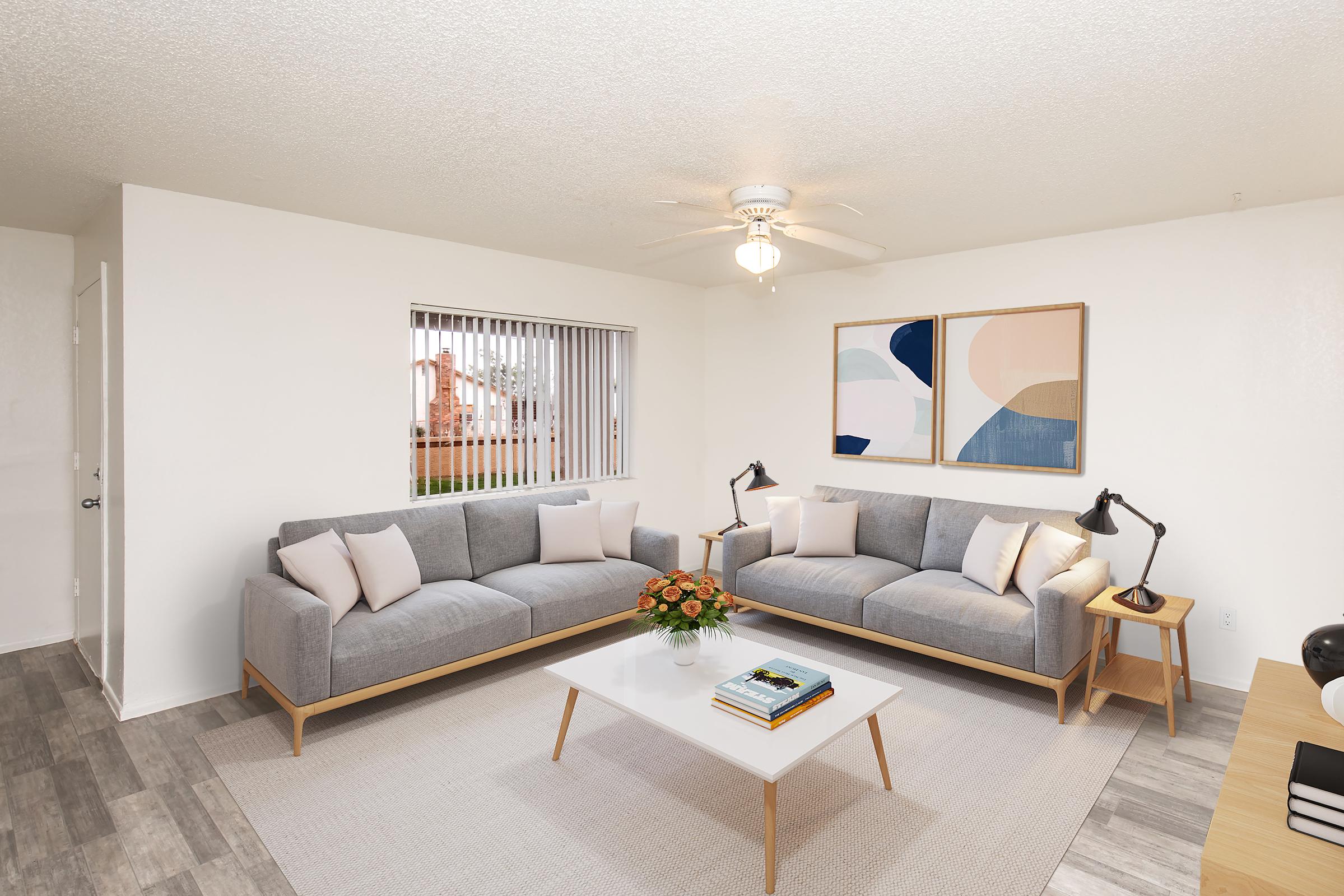 A modern living room featuring two gray sofas with light pillows, a white coffee table, and a rug. There are two stylish lamps on either side of the sofas, and a vase of orange flowers on the table. The walls are white, with a large window showcasing a view outside. Two abstract art pieces hang on the wall.