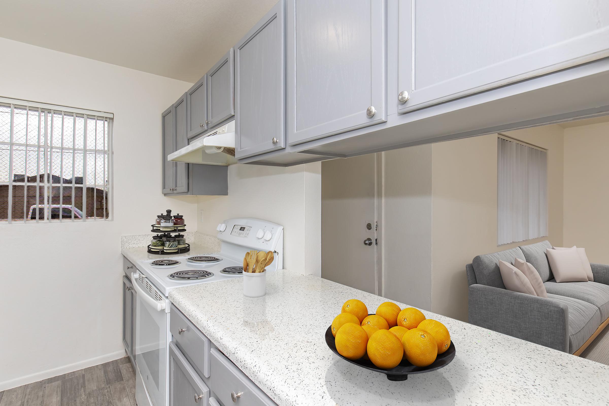 A modern kitchen featuring light gray cabinets and a white countertop. In the foreground, a bowl of oranges sits on the counter. The background includes a stove, a window with a grid pattern, and a living area visible through an open doorway, showcasing a gray sofa and light-colored walls.