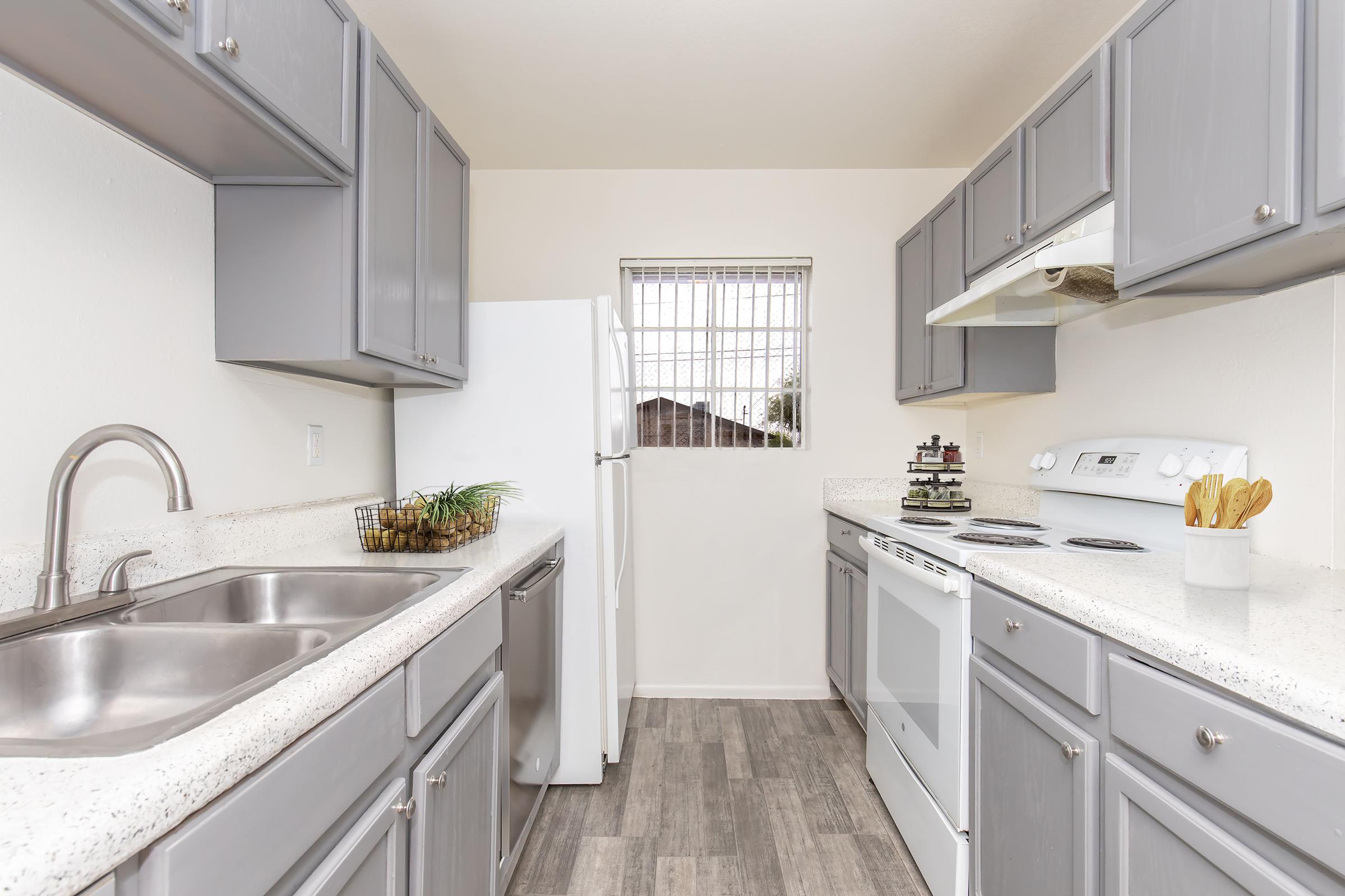 A modern kitchen featuring gray cabinets, a white refrigerator, and an electric stove. The countertops are light-colored with a speckled design, and there's a window with grid panes letting in natural light. The sink has a brushed metal faucet, and utensils are neatly arranged in a holder.