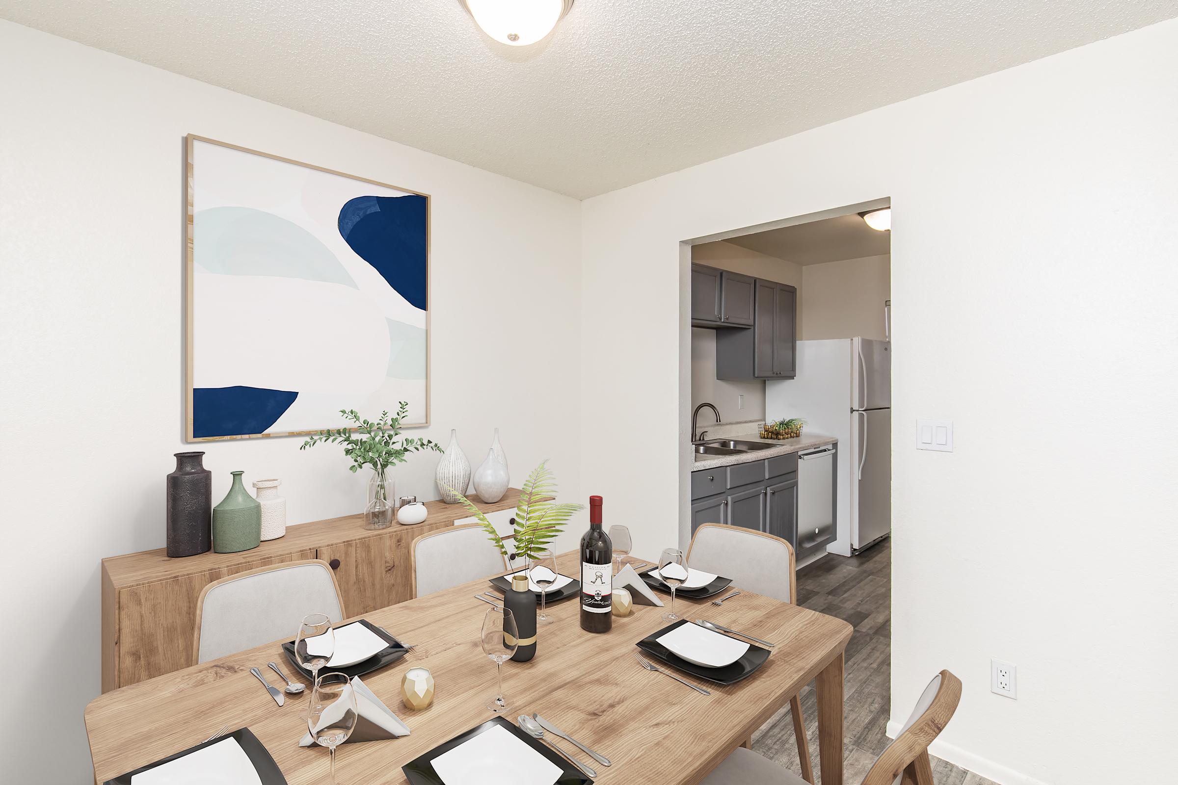 A bright and modern dining area featuring a wooden dining table set with plates, glasses, and cutlery. A large abstract painting hangs on the wall, and a sideboard holds decorative vases and a bottle of wine. An open doorway leads to a kitchen with gray cabinetry visible in the background.