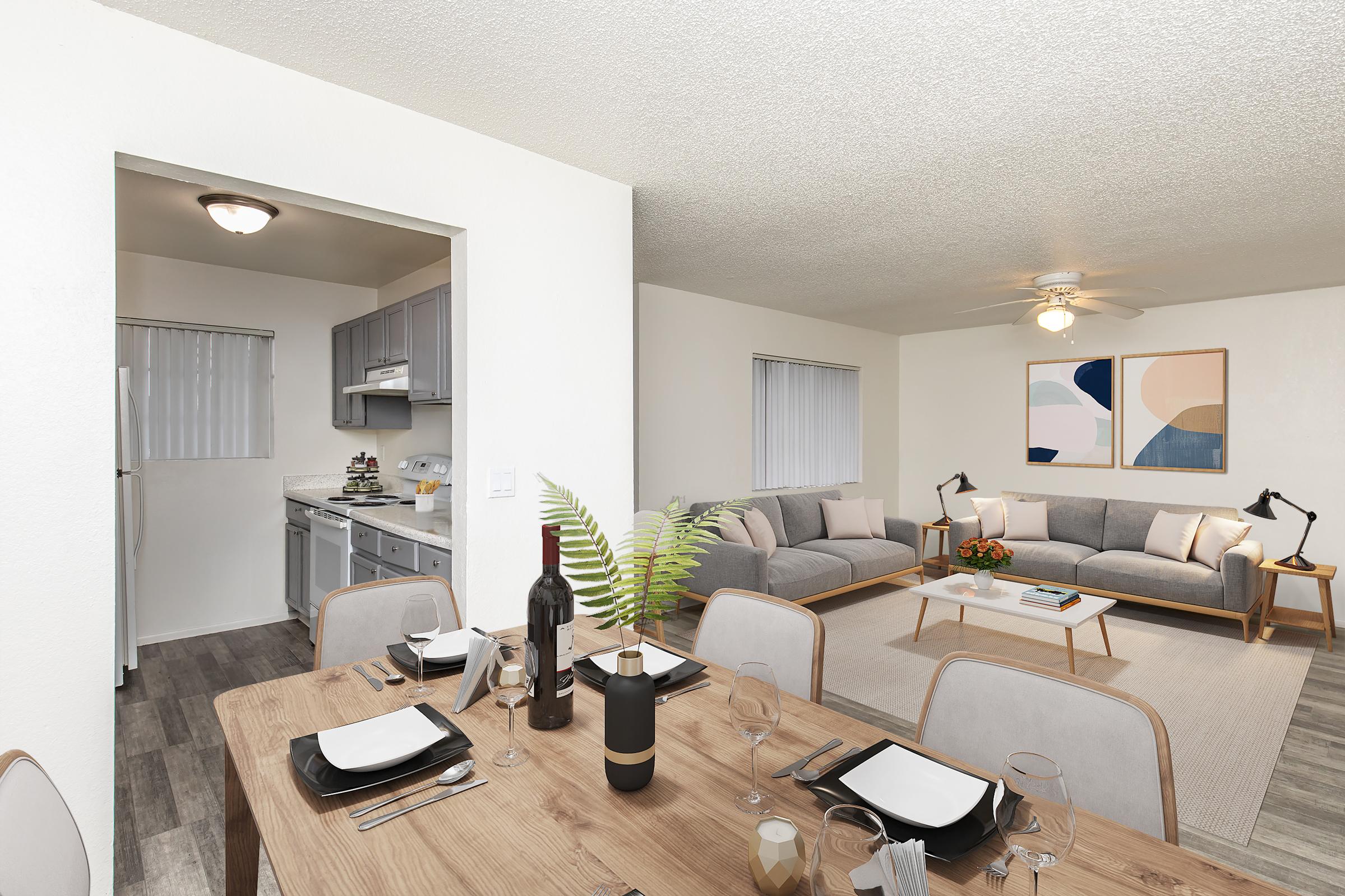 A modern living and dining area featuring a wooden dining table set for four, with wine and glasses. The adjacent living room includes two gray sofas and a coffee table, complemented by abstract wall art. A kitchen area is visible in the background, with light-colored cabinets and appliances.