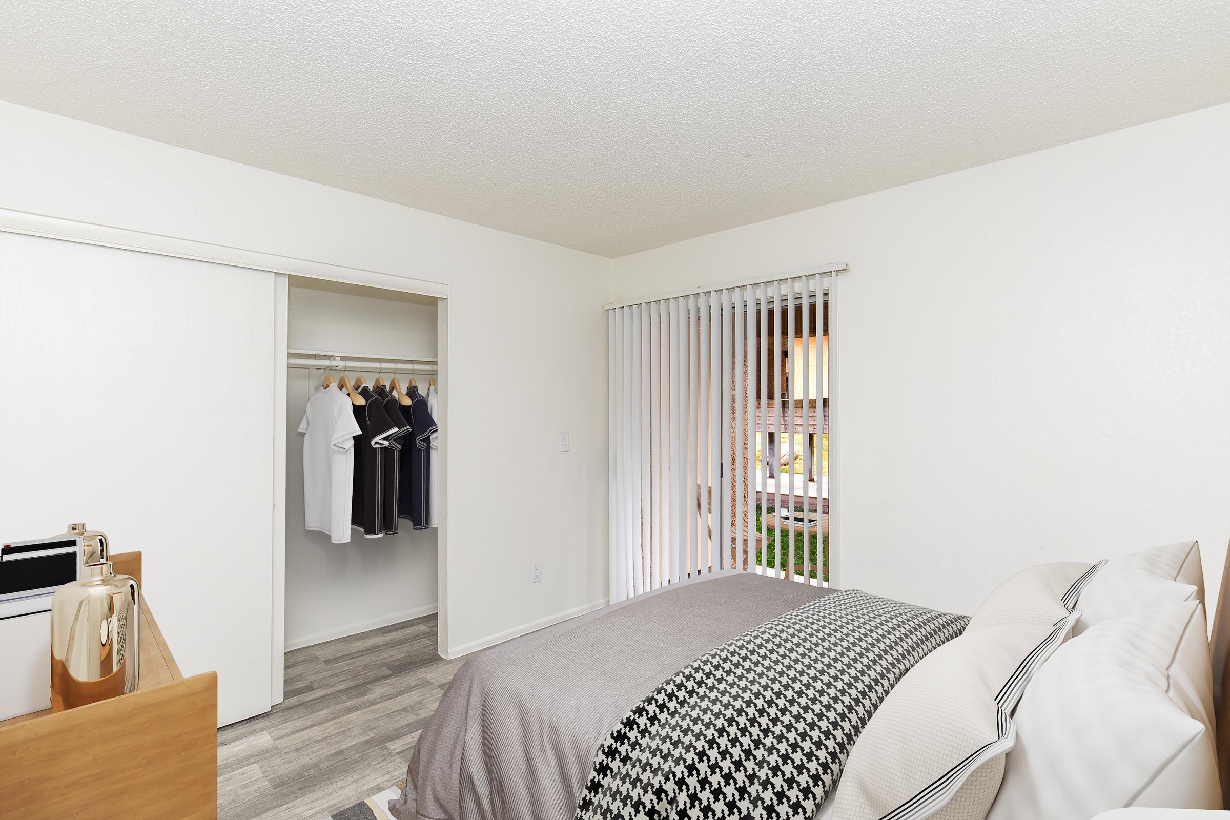 A well-lit bedroom featuring a neatly made bed with gray and white bedding. A closet with hanging clothes is visible on the left, and sliding vertical blinds allow natural light from a window. The floor has light-colored wood-like planks, creating a clean and modern aesthetic.