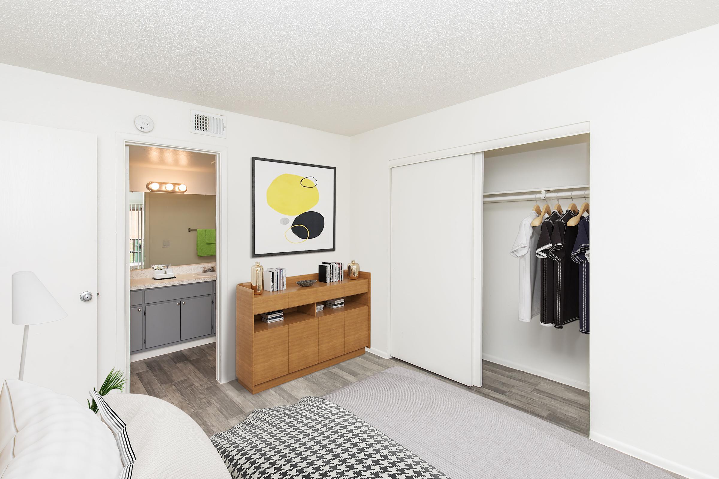 A modern bedroom featuring a bed with a patterned throw blanket, a wooden dresser displaying decorative items, and an open closet with hanging clothes. A bathroom is visible in the background, and a framed abstract artwork adorns the wall. The room has neutral walls and stylish flooring.