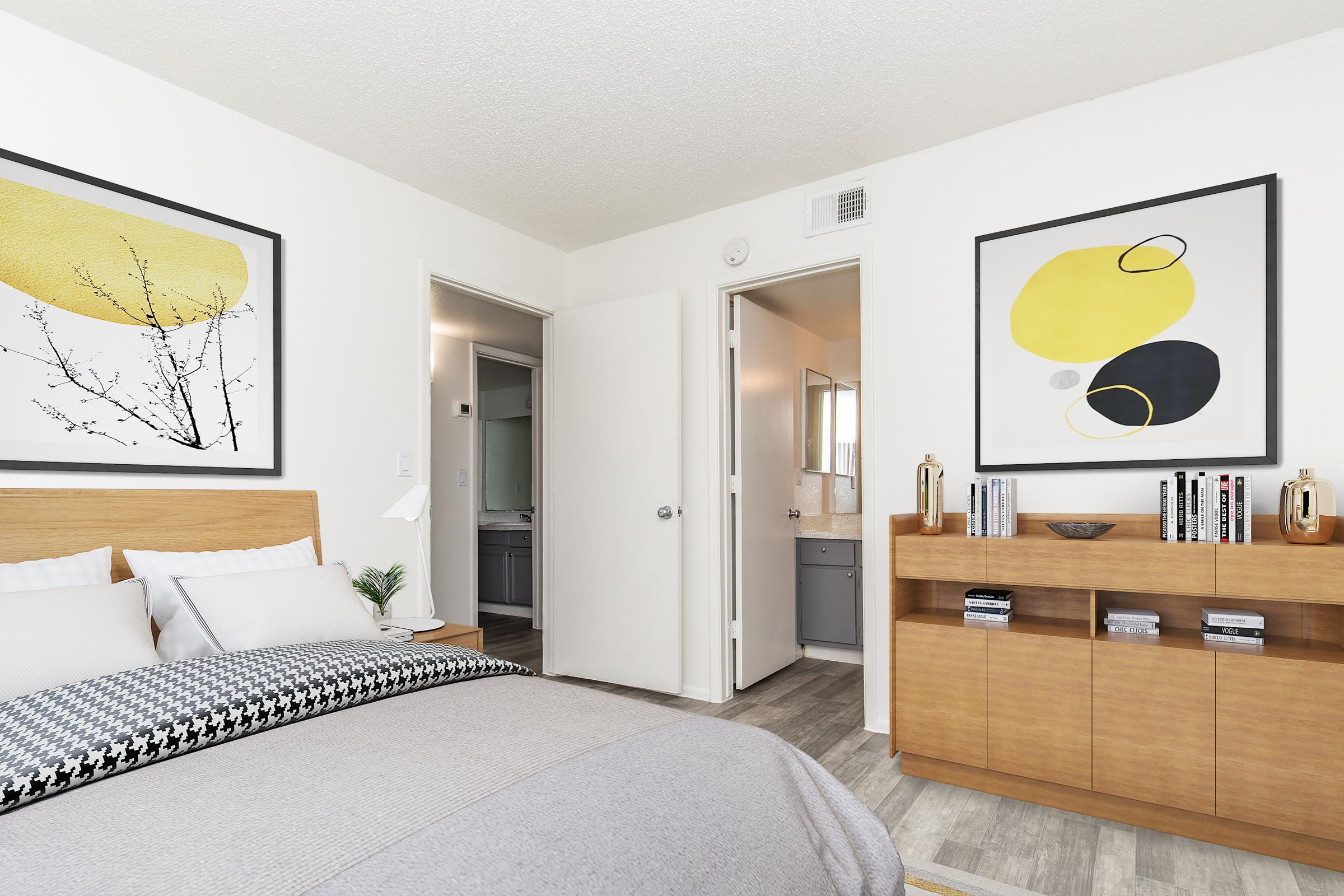 A bright, modern bedroom featuring a large bed with gray and white bedding, two decorative wall art pieces above the headboard, and a wooden side table. A door leads to a bathroom, while a wooden cabinet on the opposite wall holds books and decorative items. The room has neutral-colored walls and a polished floor.