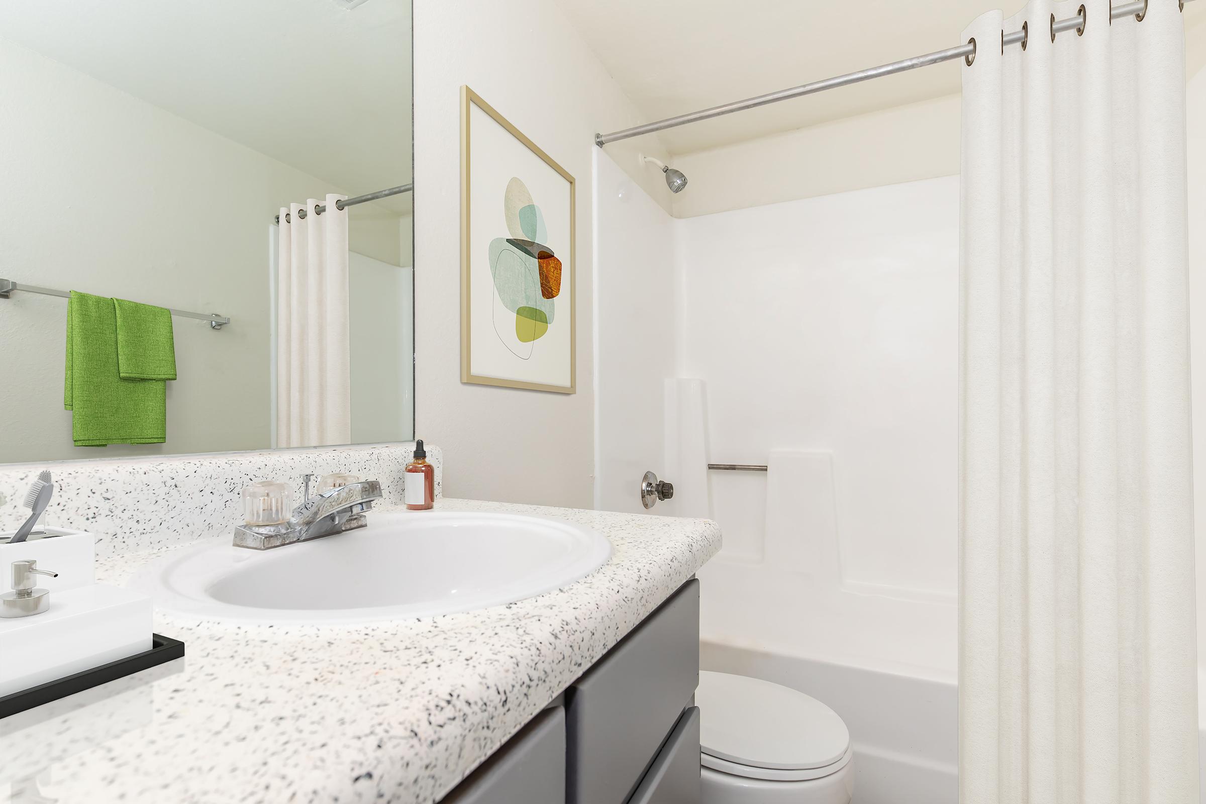 A clean bathroom featuring a light-colored countertop with a sink, a white bathtub with a shower curtain, and a decorative framed artwork on the wall. Green towels are neatly hung on the rack, adding a pop of color to the neutral decor.