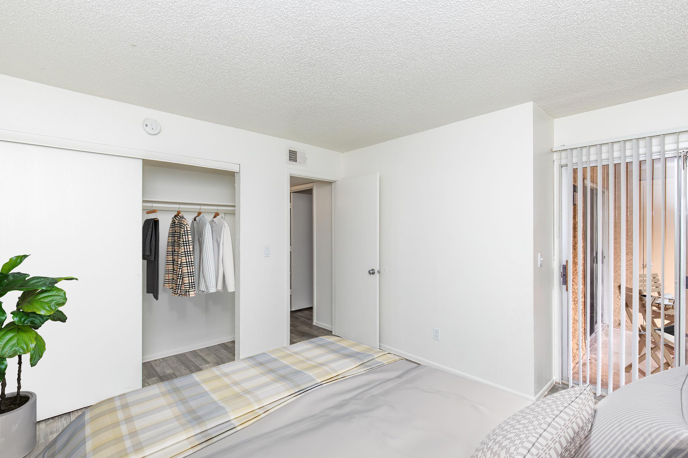 A bright and spacious bedroom featuring a neatly made bed with plaid bedding. There is a closet with several items hanging inside, including a coat and a patterned shirt. The room has light-colored walls, a potted plant in the foreground, and sliding glass doors leading to a balcony or patio area.
