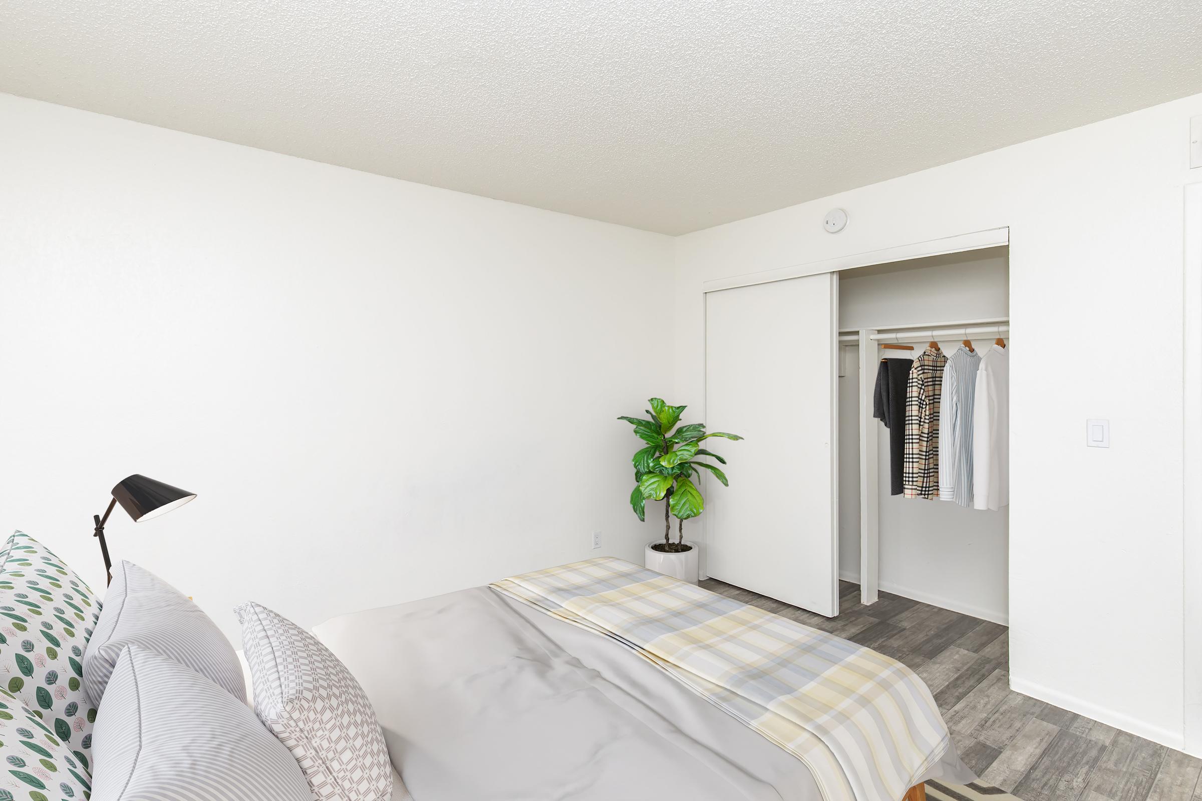 A bright, modern bedroom featuring a neatly made bed with patterned pillows and a light-colored blanket. To the right, there is a potted plant beside a closet with sliding doors, showcasing hanging clothes inside. The walls are painted white and the flooring is composed of wood-like tiles.