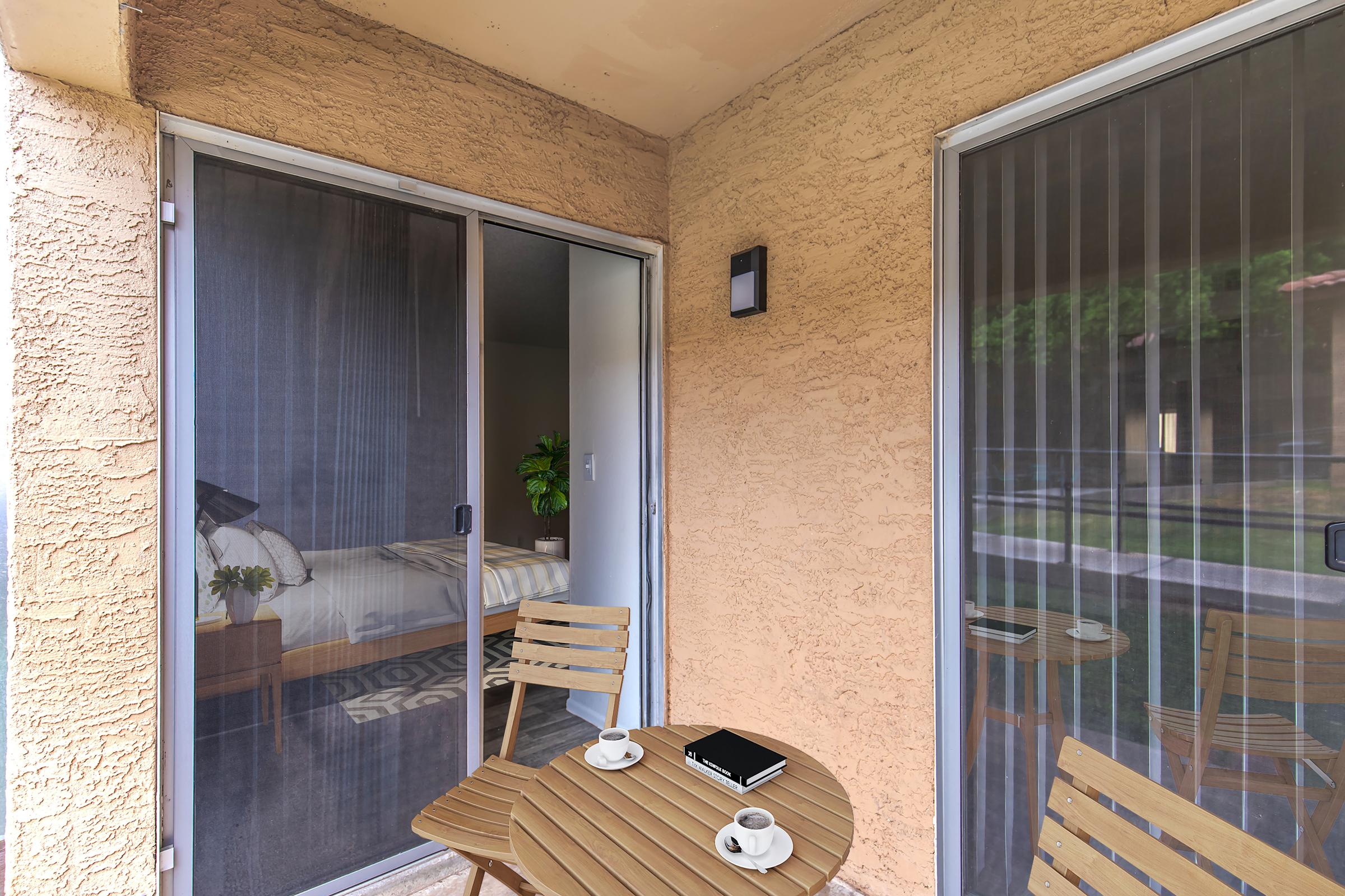A cozy balcony with two wooden chairs and a small round table, showcasing two coffee cups and a notebook. The background features sliding glass doors opening into a bedroom with a neatly made bed, and the exterior wall has a textured finish. Natural light streams onto the outdoor space.