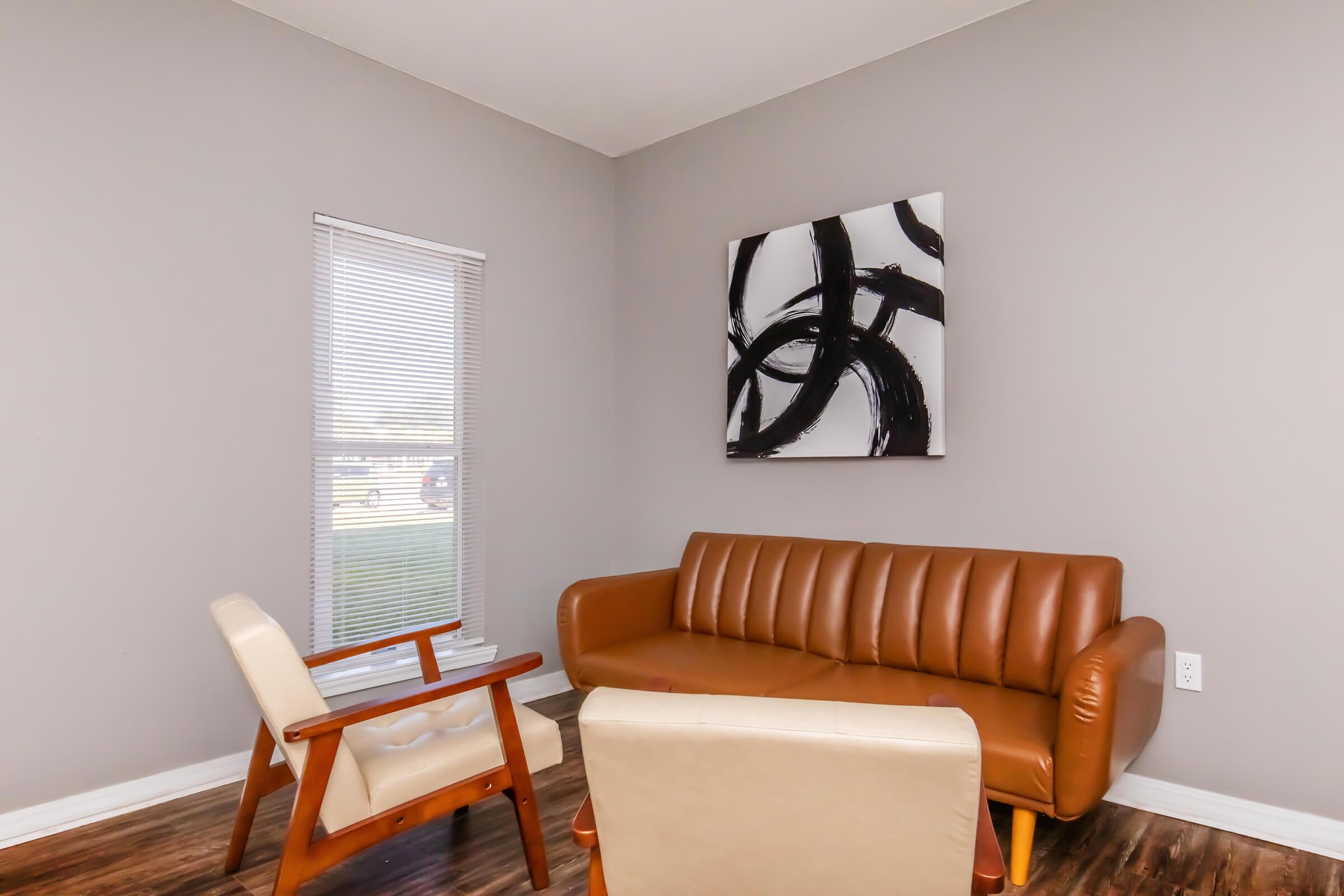 A cozy living room featuring a brown leather sofa and an armchair, set against gray walls. A piece of abstract black artwork hangs above the sofa, and a window with blinds allows natural light to enter the space. The floor is wooden, creating a warm and inviting atmosphere.