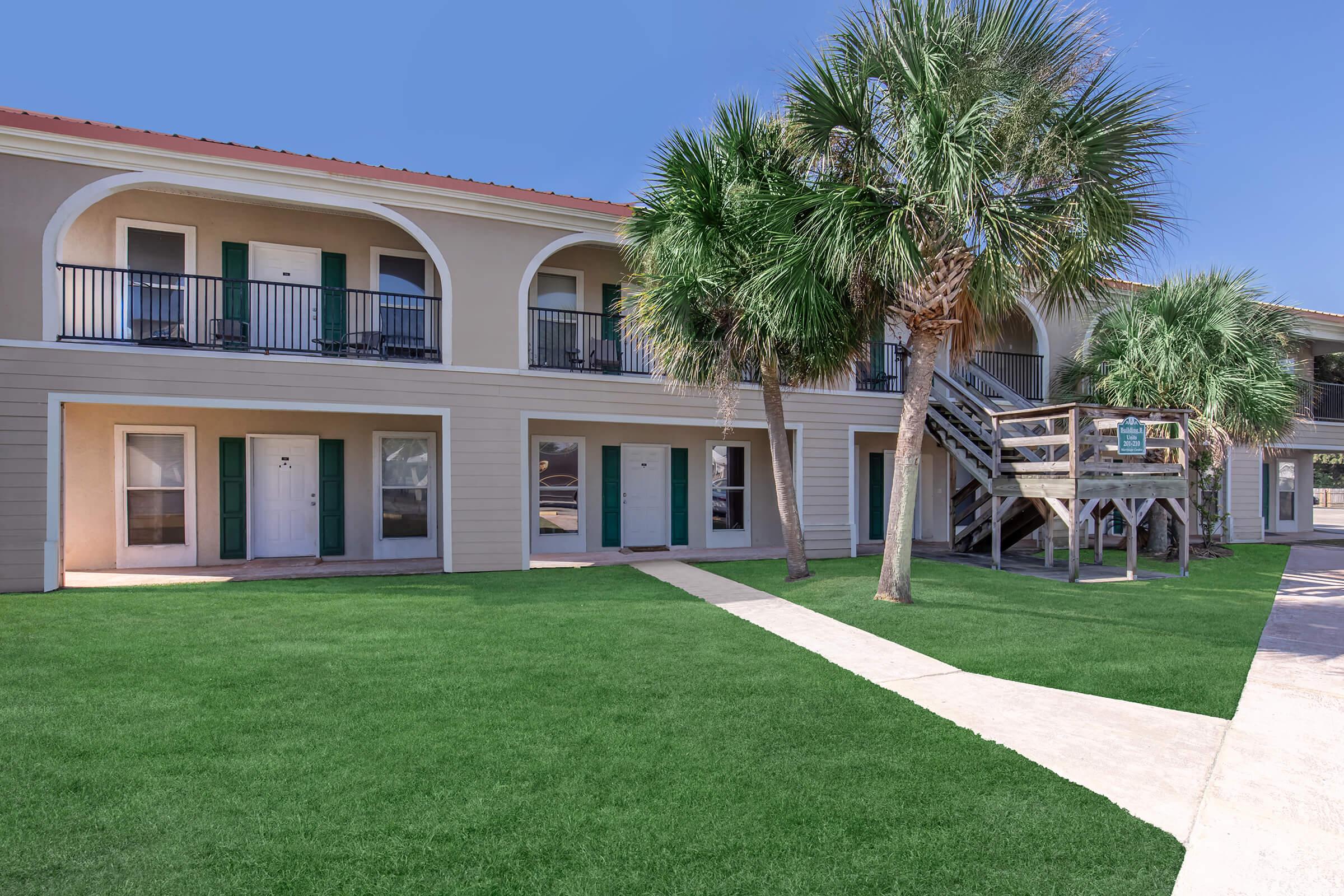 A view of a two-story motel with a sandy beige exterior, featuring green doors and balconies. Lush palm trees surround the building, and a grassy area leads to a pathway. The sky is clear and blue, providing a pleasant atmosphere. The design suggests a tropical or beachside location.