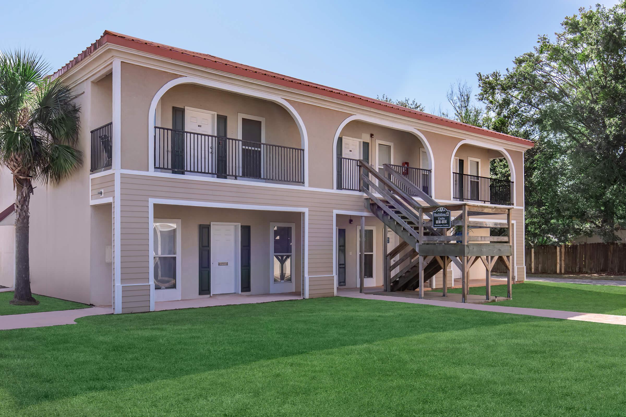 Two-story building with a beige exterior and archway balconies. The ground level features dark wooden stairs leading to the upper level. Lush green lawn surrounds the building, and palm trees are visible nearby. sunlight illuminates the scene, creating a bright and inviting atmosphere.