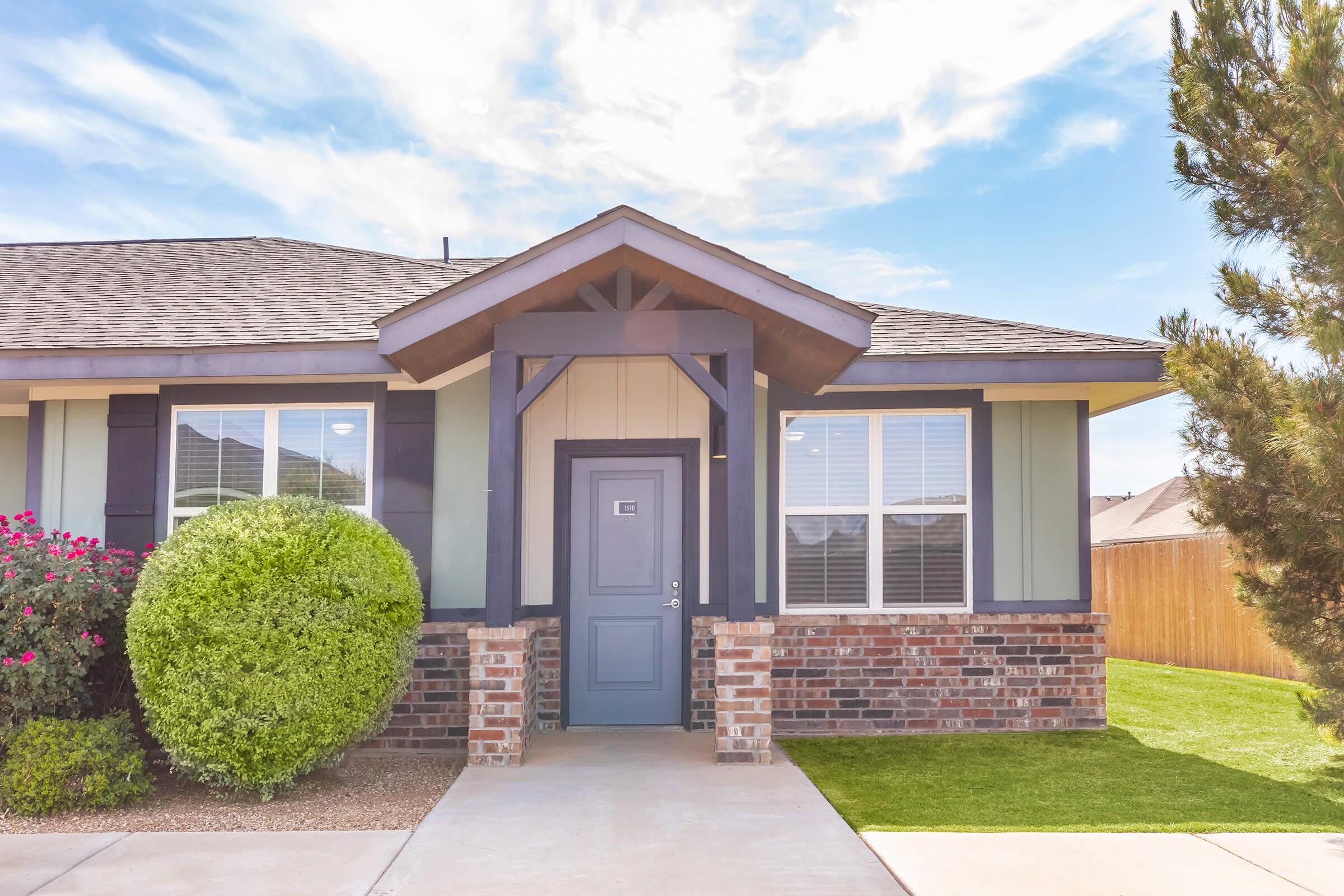A well-maintained single-story house with a gray door and decorative wood accents. The house features multiple windows, a manicured lawn, and a rounded shrub. Vibrant flowers are visible near the entrance, and the sky is clear with soft, scattered clouds.