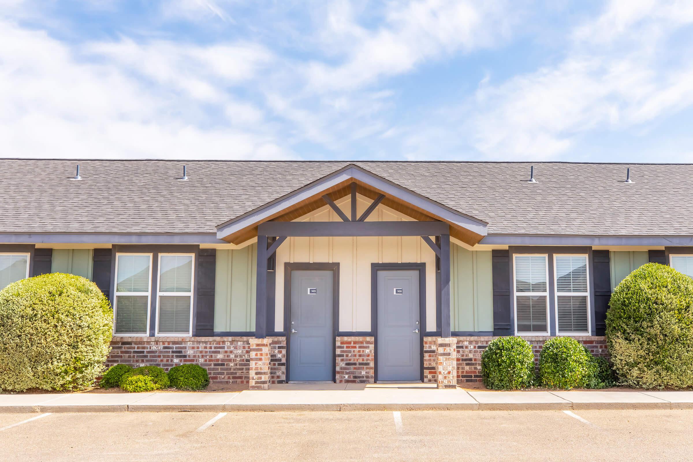 Exterior view of a single-story residential building featuring two entrance doors. The structure has a peaked roof and is surrounded by well-maintained bushes. A clear blue sky is visible in the background, providing a bright and inviting atmosphere. The setting appears to be in a suburban area.